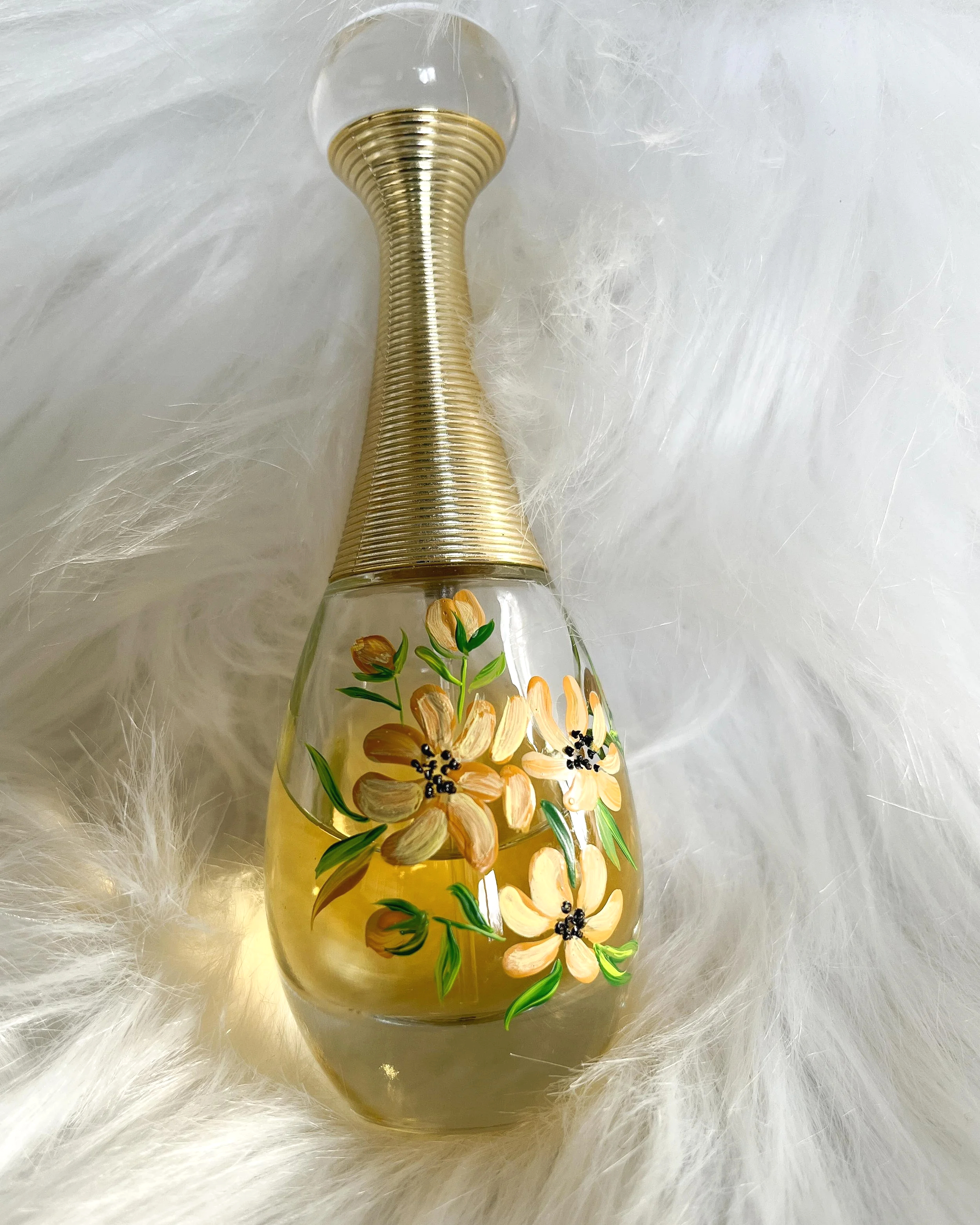 Decorative glass perfume bottle with painted yellow and orange flowers and green leaves, silver ribbed neck, lying on white furry surface.