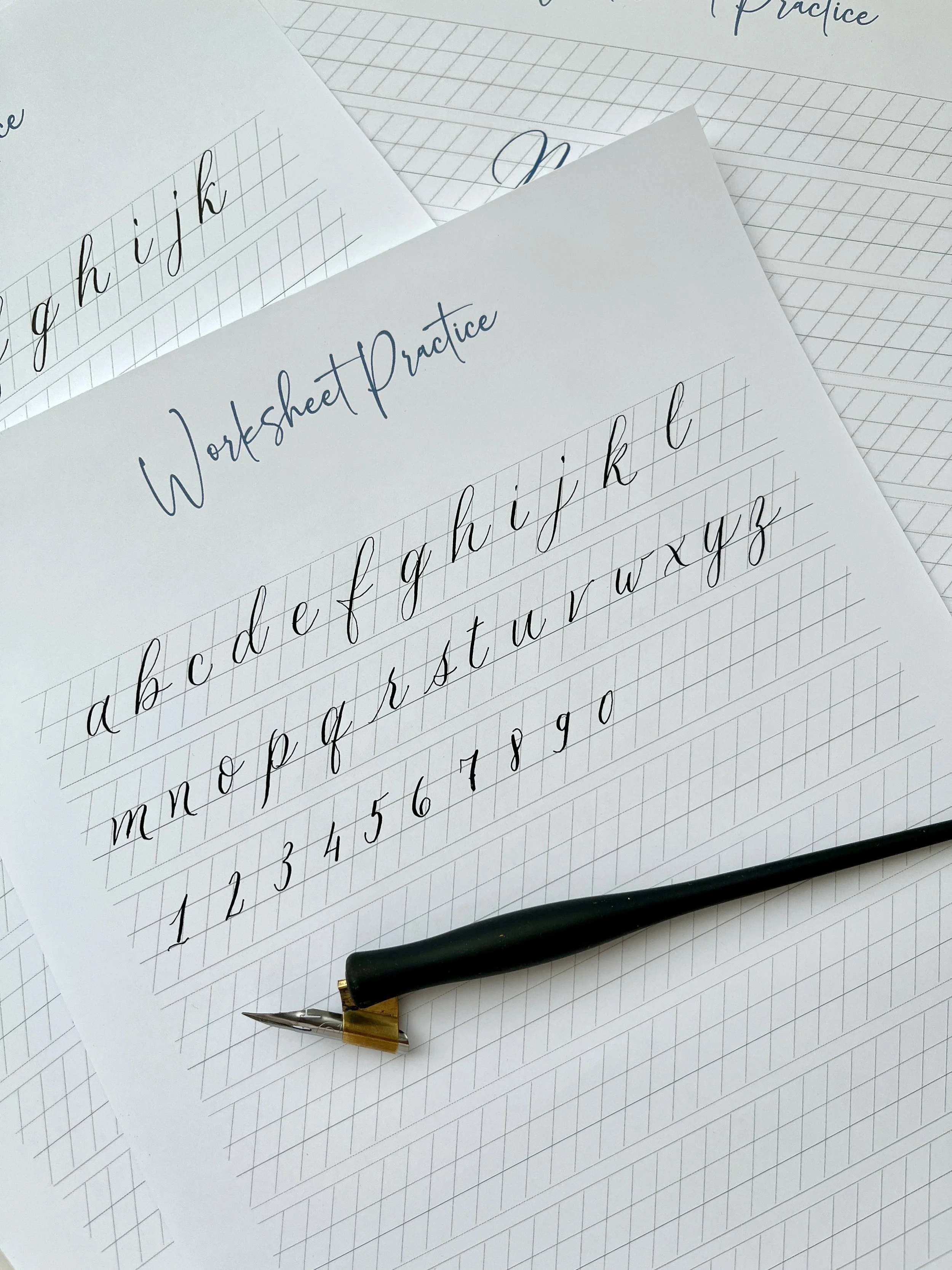 Sheets of paper with cursive handwriting practice including alphabet letters and numbers, along with a dip pen.