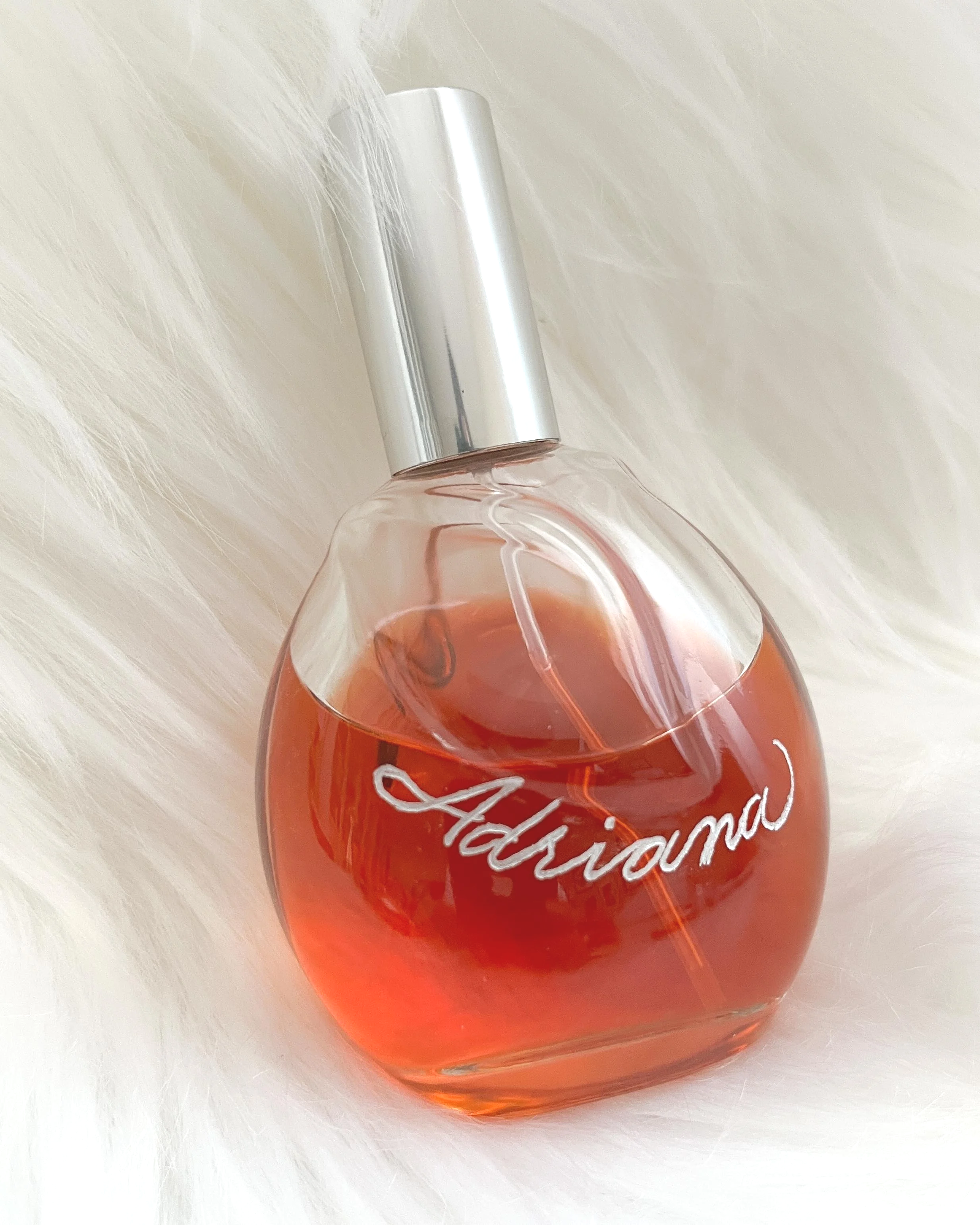 A glass perfume bottle labeled 'Adriana' with pink liquid inside, resting on a soft white furry surface.