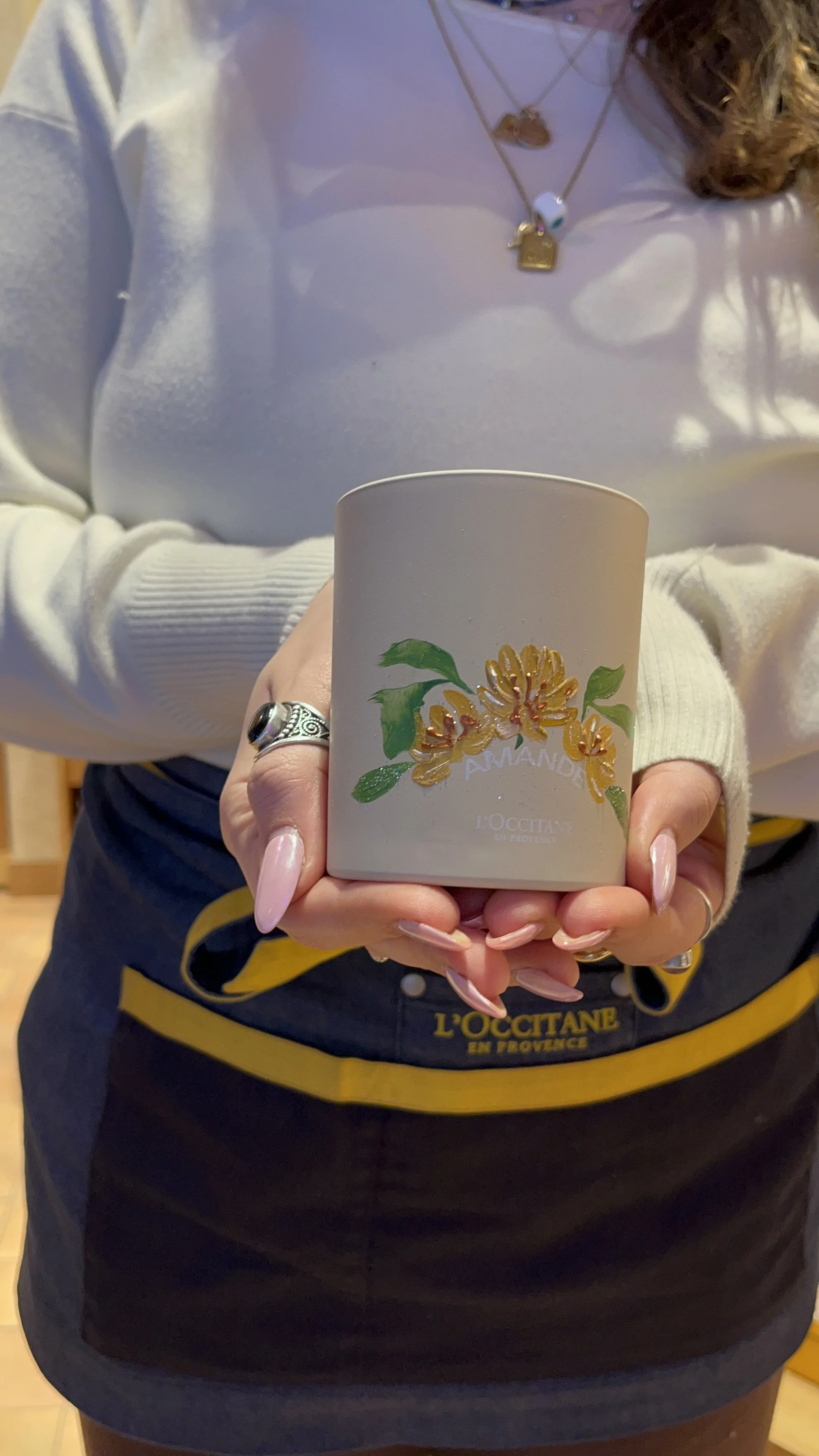 A person holding a white candle with a floral design and the words 'Amand' on it. The person is wearing a white sweater, jewelry including rings and layered necklaces, and a black apron with yellow writing. The background appears to be a wooden interior.
