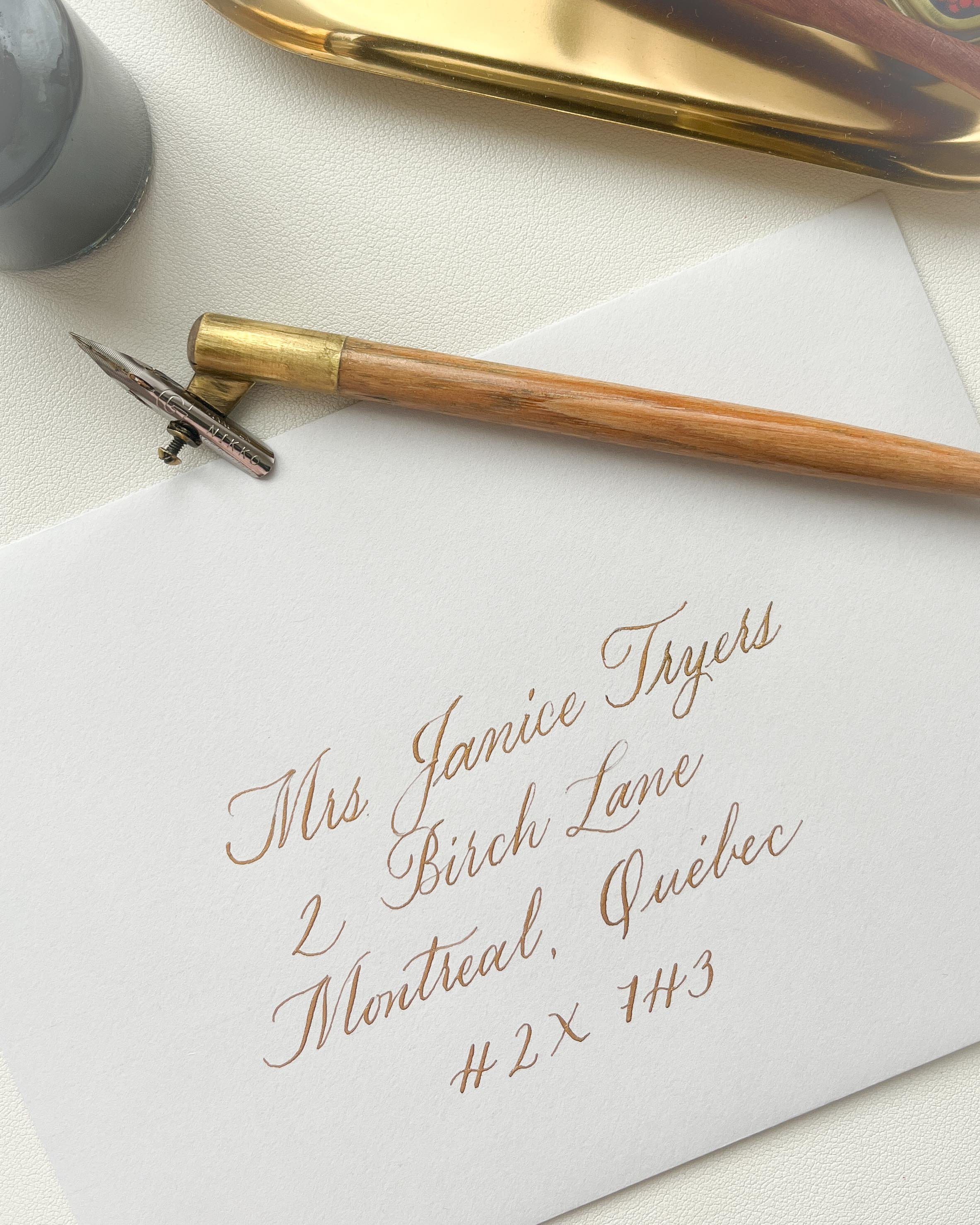 A calligraphy pen with a wooden handle and a metal nib, placed on a white paper with elegant gold calligraphy text. The paper contains an address written in calligraphy, which reads: "Mrs. Janice Tryers, 2 Birch Lane, Montreal, Quebec, #2X 1H3." There is also a gold tray and a small dark container partially visible at the top of the image.