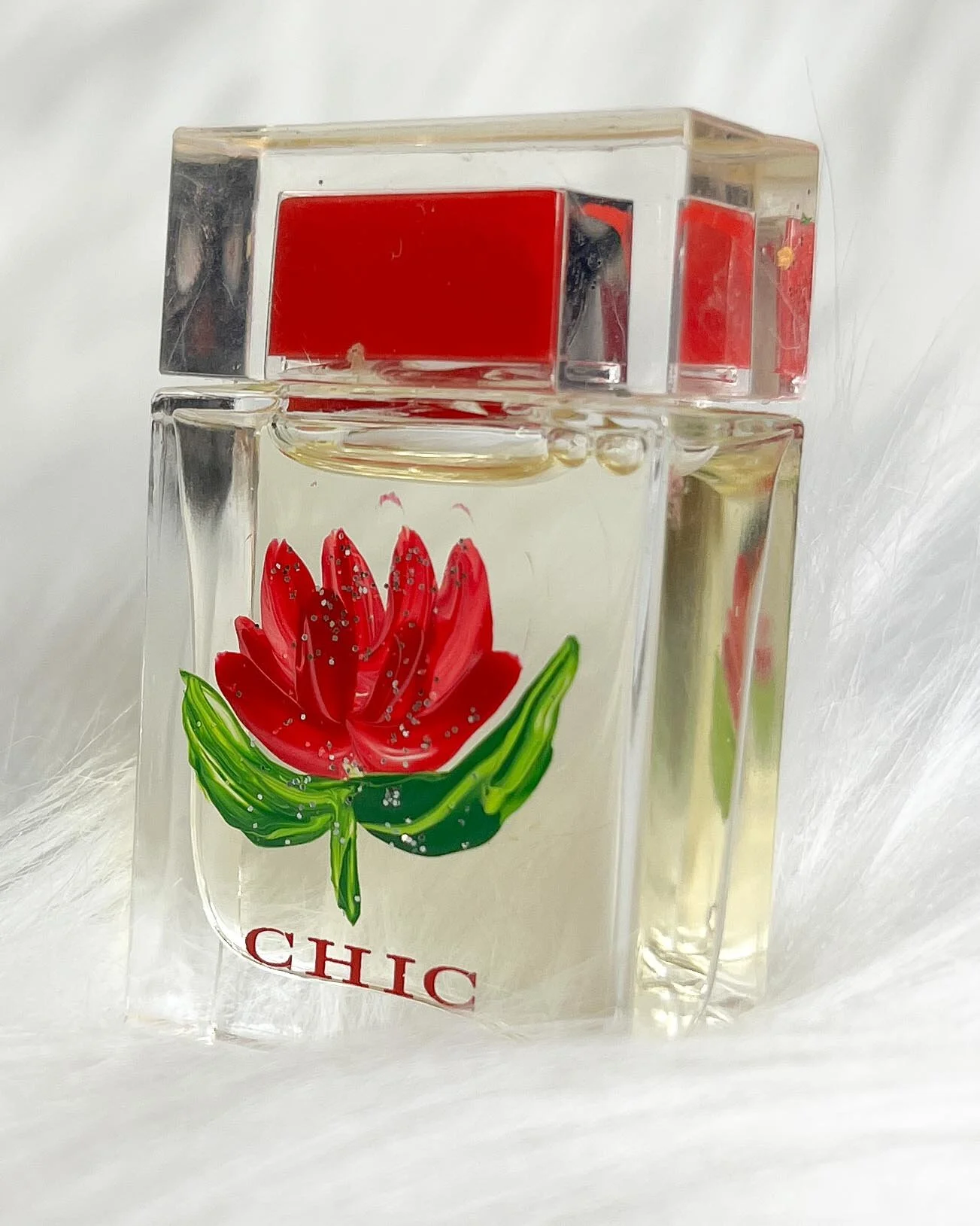 A clear glass perfume bottle with a red flower design and the word "CHIC" printed on it, topped with a transparent cap with a red accent.