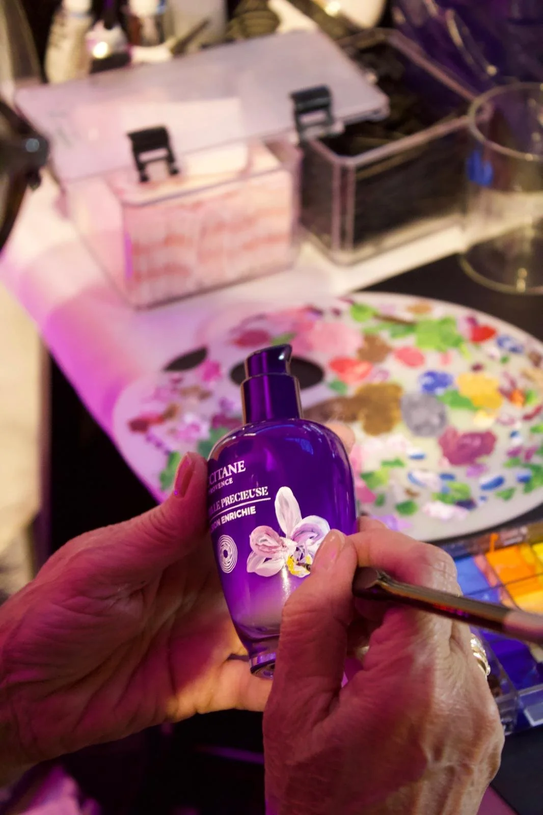 Person holding a purple bottle of L'Occitane floral perfume in front of a colorful palette and containers of paint on a table.