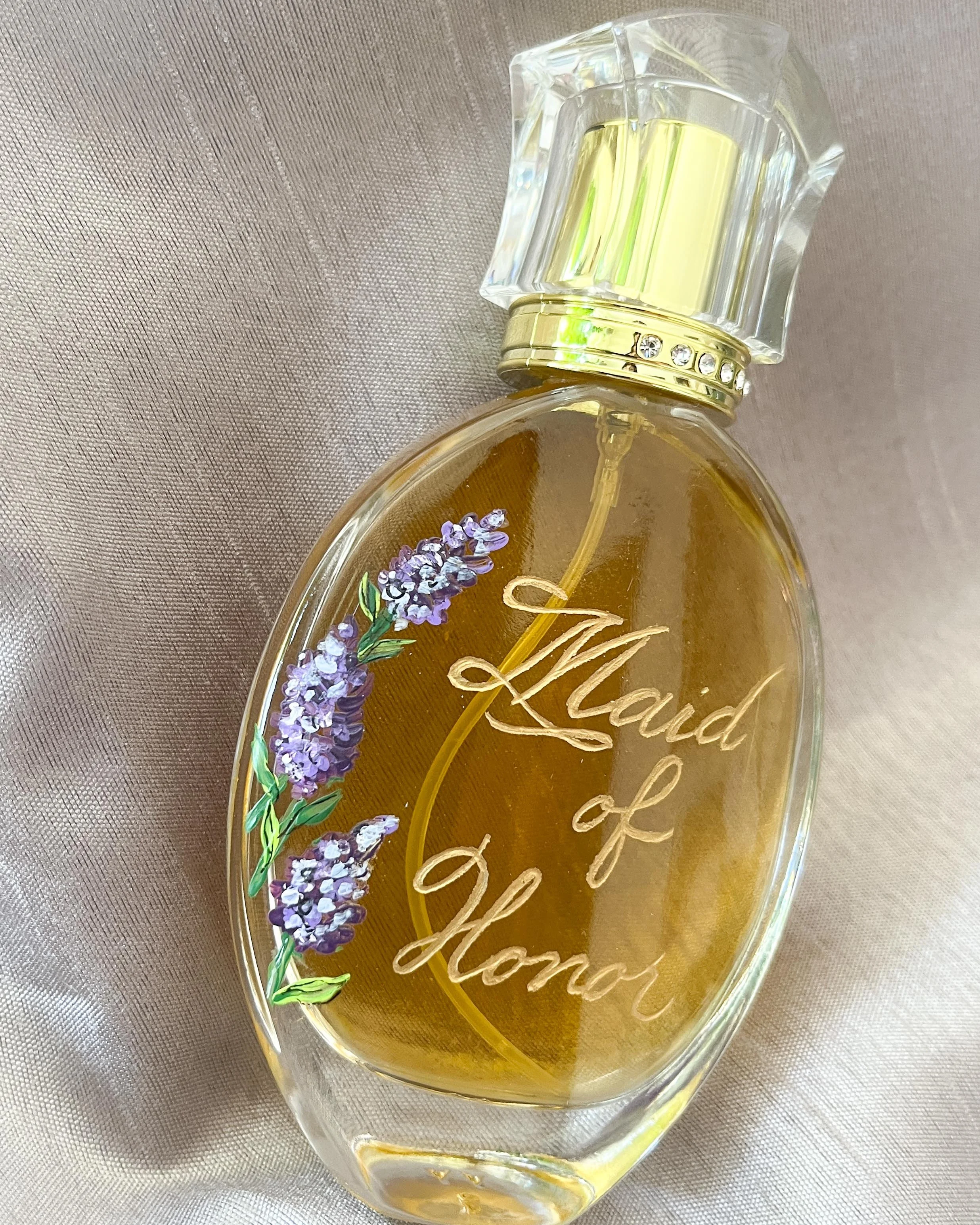A clear perfume bottle with gold accents and a lavender flower design, labeled "Laid of Honor," resting on a fabric surface.