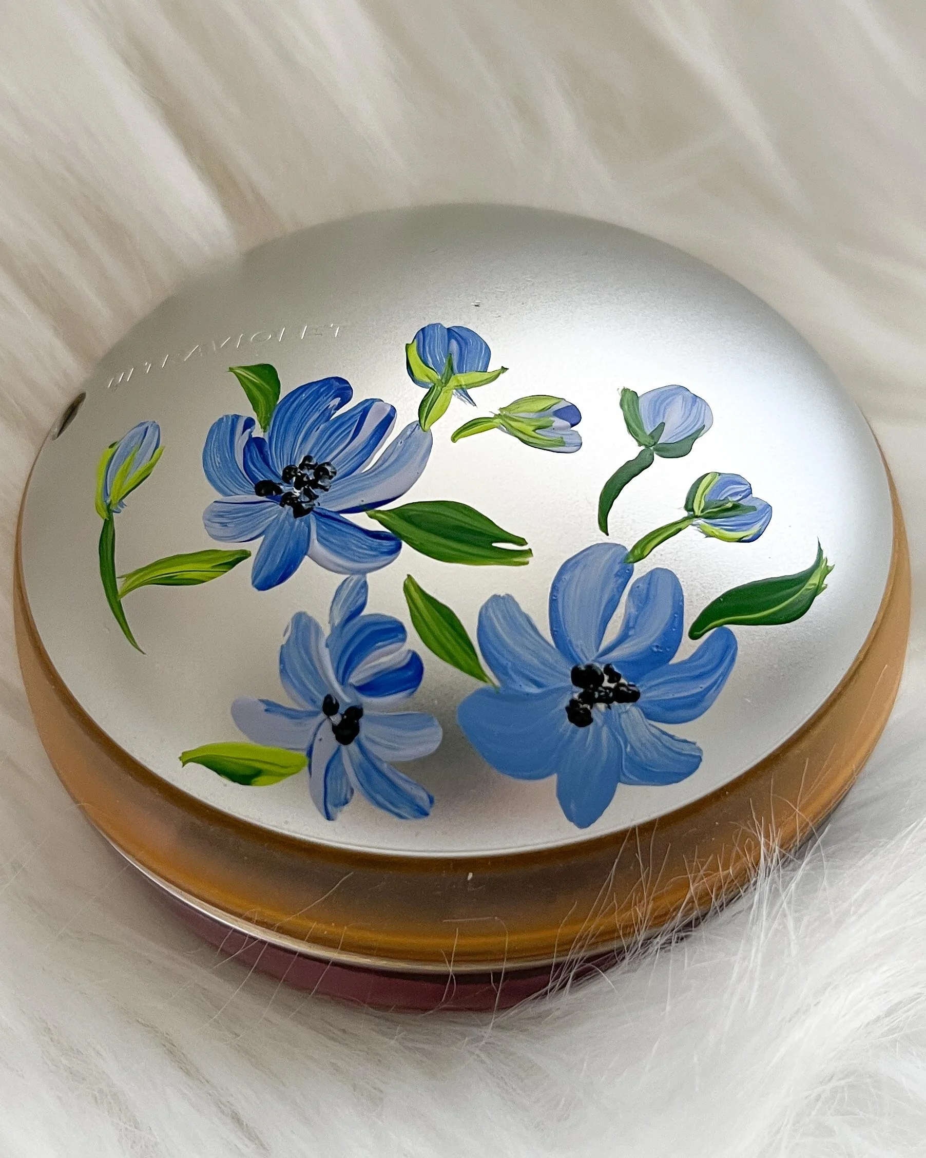 Spherical mirror with painted blue flowers and green leaves, placed on a white furry surface.