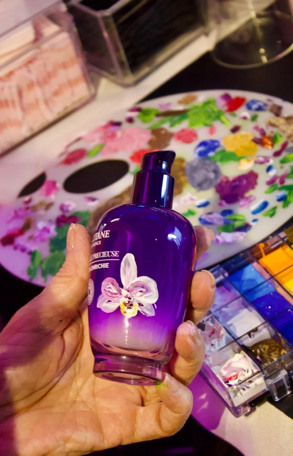 A hand holding a purple bottle of fragrance with a white orchid on the label. In the background, there is a colorful artist's palette with paint splotches, and various makeup supplies, including a container with brushes and a drawer unit with organized makeup products.