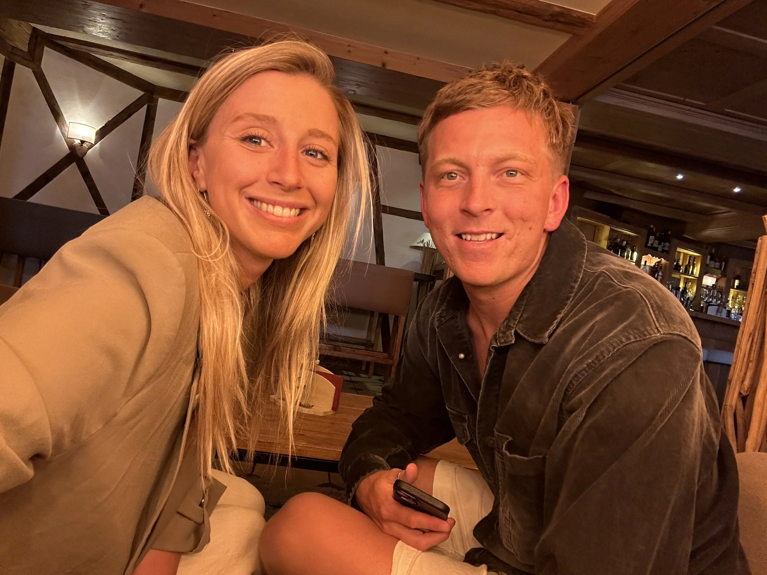 A smiling woman with long blonde hair and a man with short light brown hair sitting at a table in a restaurant or bar, with warm lighting and wooden decor in the background.