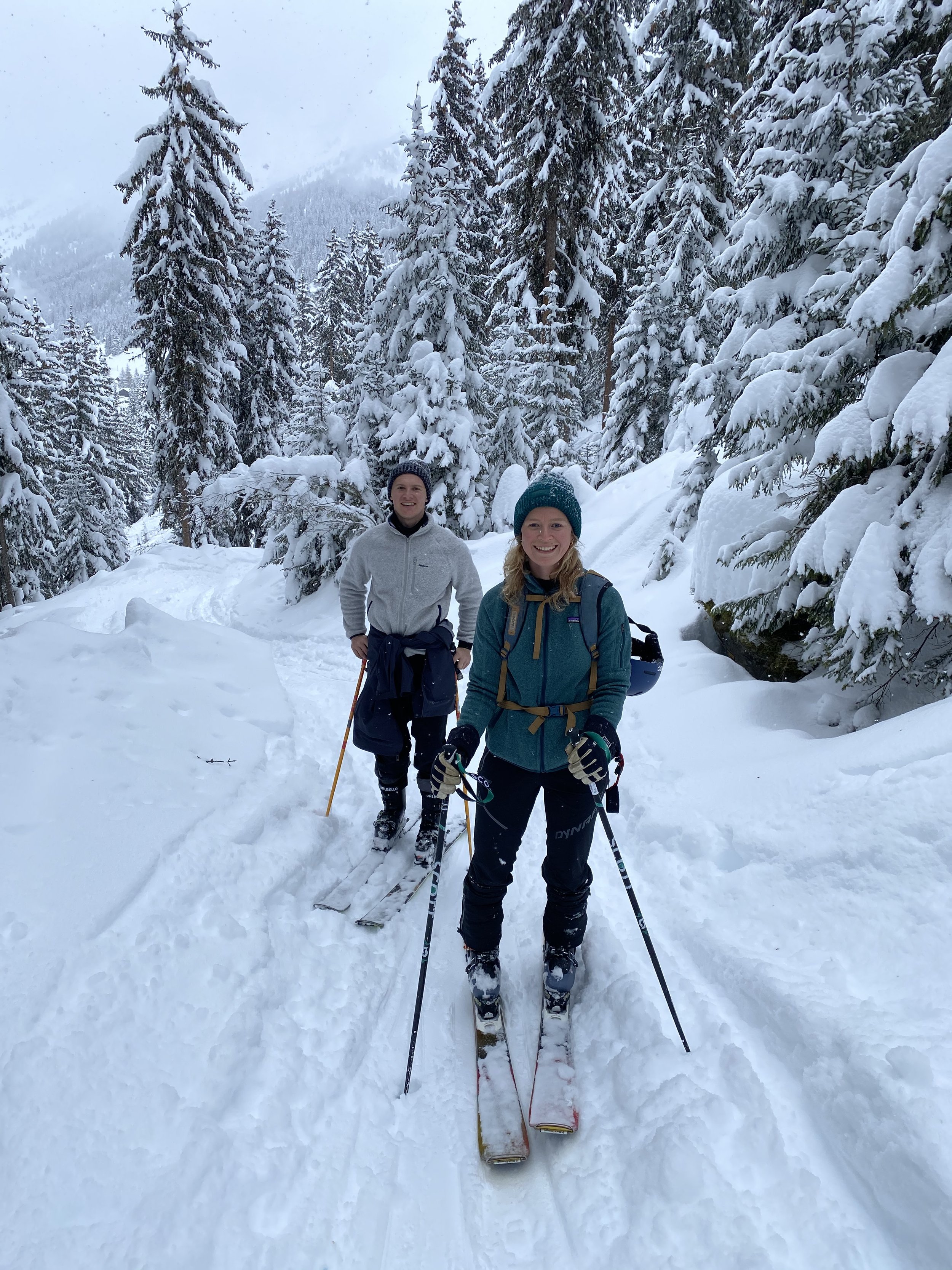 Two people skiing on a snowy trail surrounded by snow-covered trees in a mountainous area.