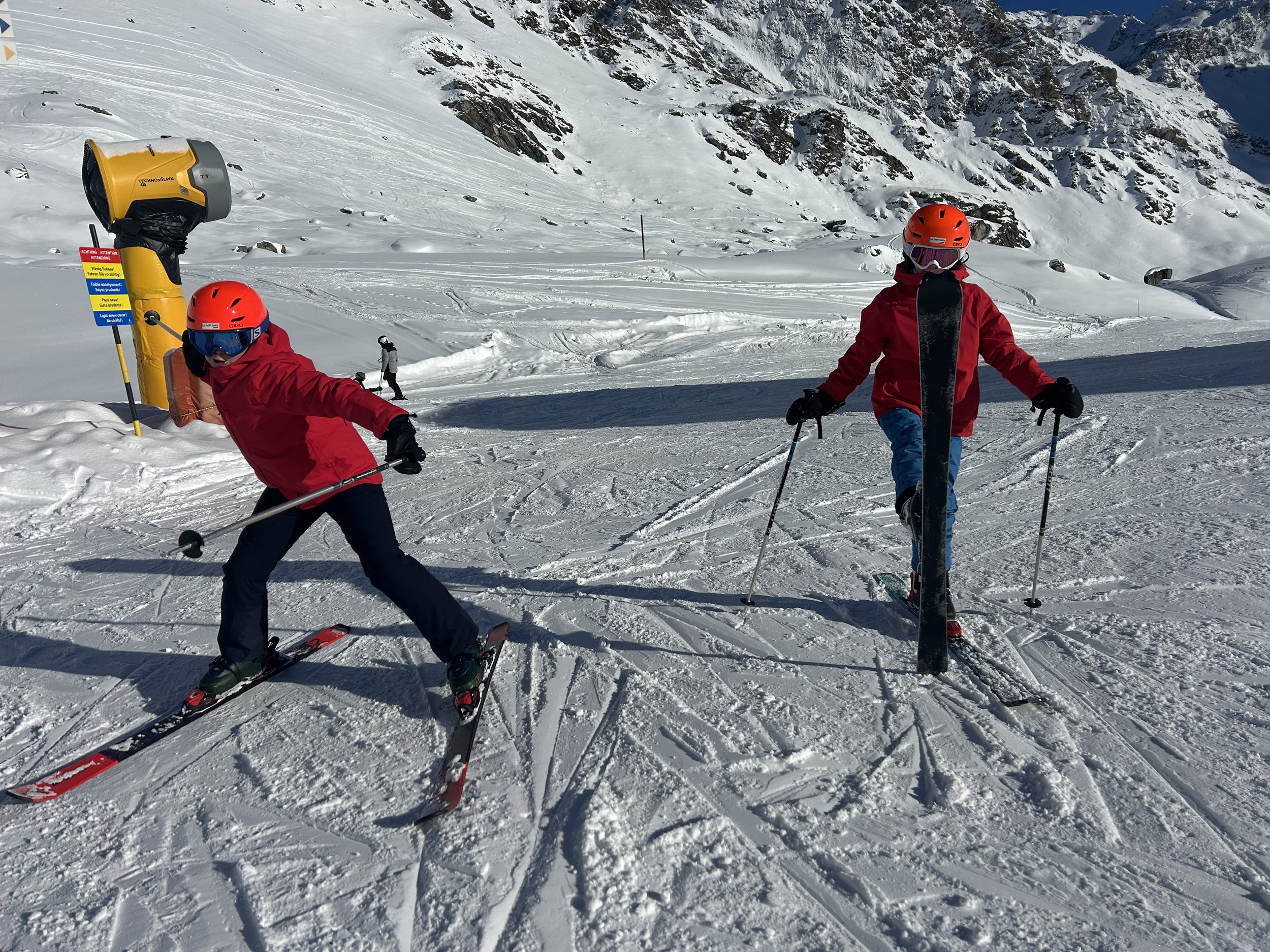 Two children in red jackets and helmets skiing on a snowy mountain slope, with ski poles in hand and a yellow ski lift in the background.