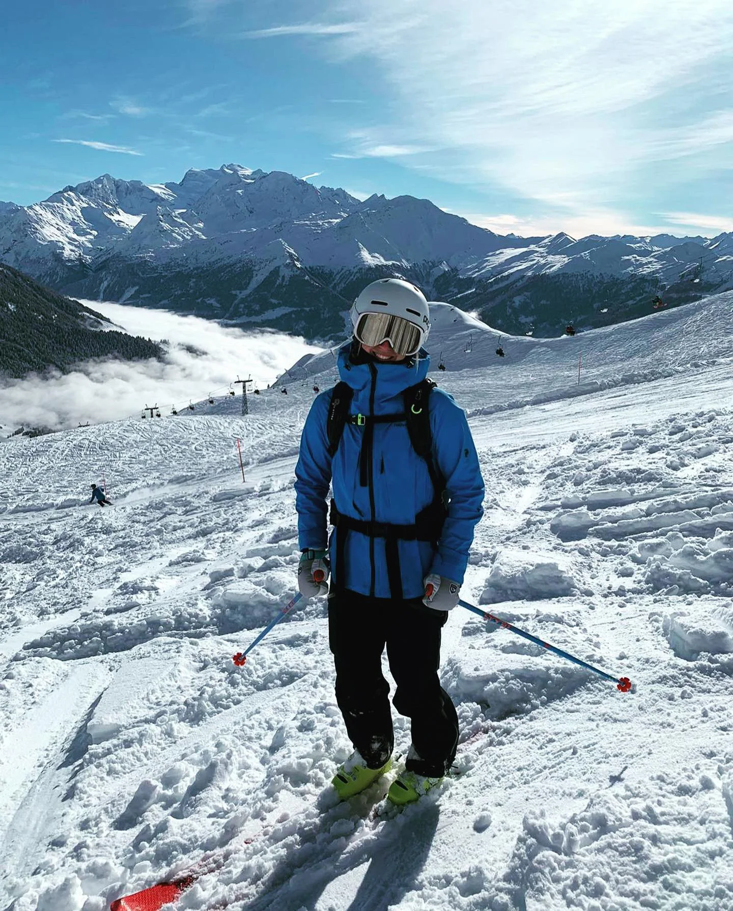 A person dressed in blue ski gear, wearing a helmet and goggles, standing on a snowy mountain slope with ski poles, surrounded by snow-covered mountains and a bright blue sky.