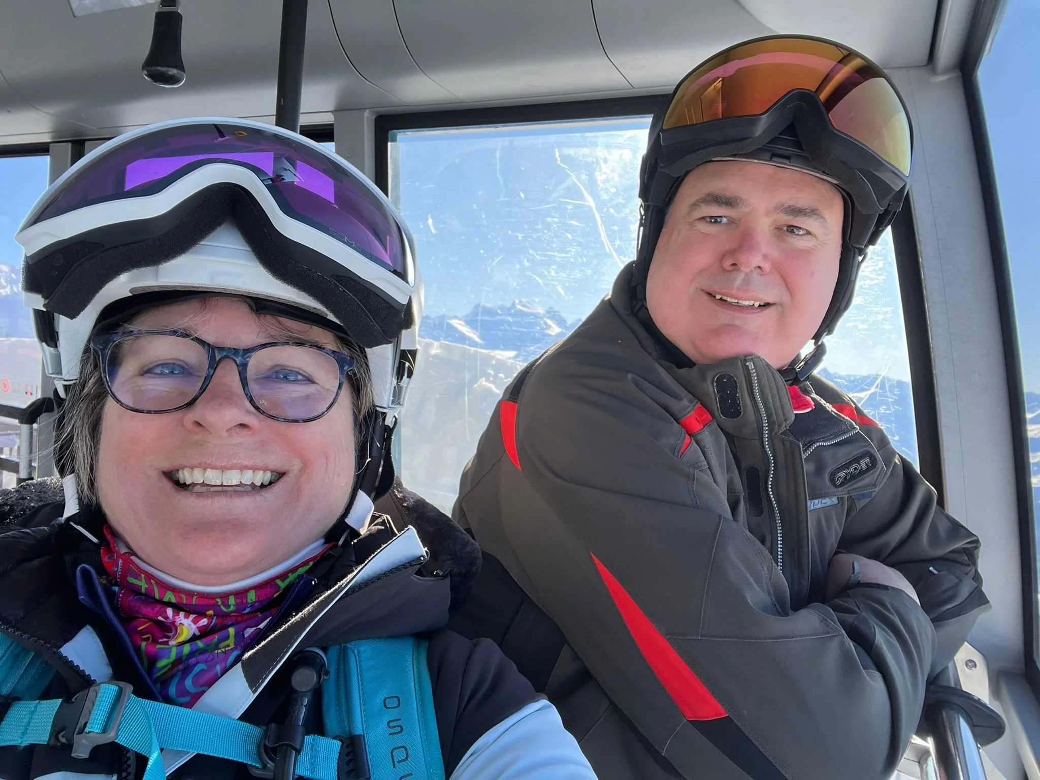 Two people in ski gear taking a selfie on a ski lift, smiling, wearing helmets and goggles.