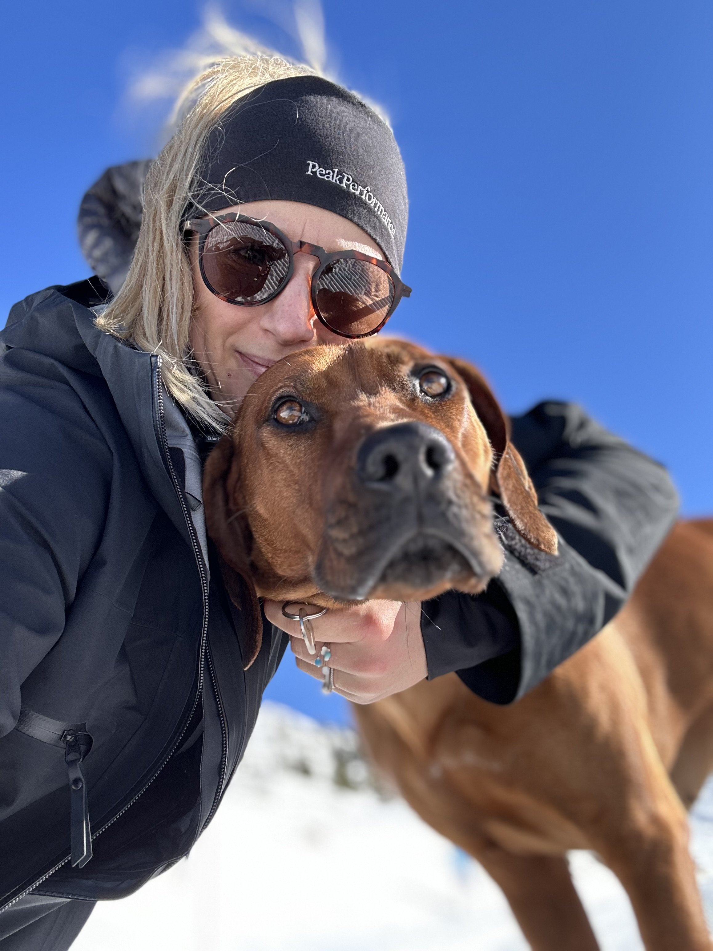 Woman wearing sunglasses and a headband taking a selfie with her brown dog outdoors on a snowy day.