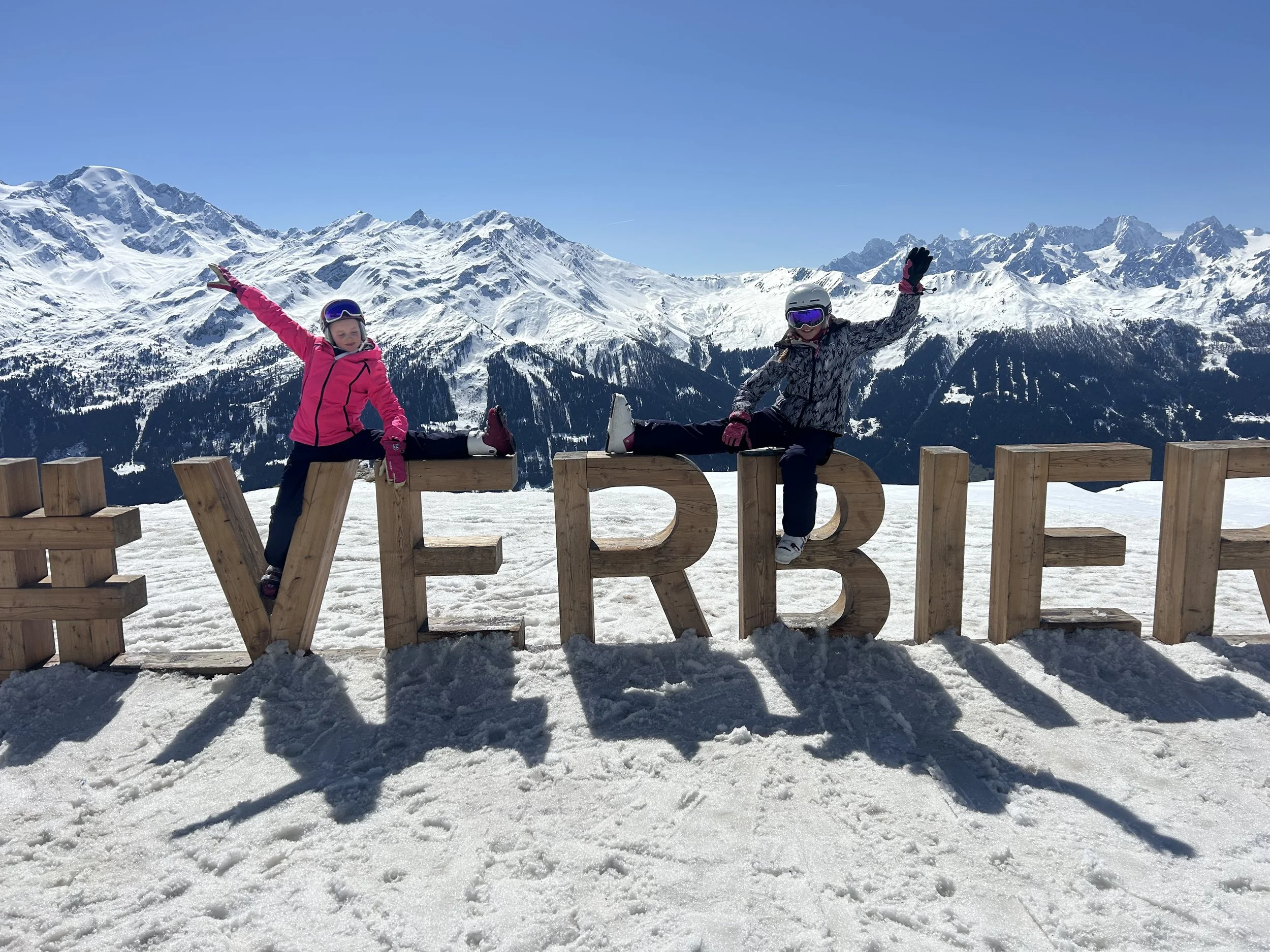 Two young girls in ski gear standing on snowy slope with skiers in background and mountain peaks in distance.