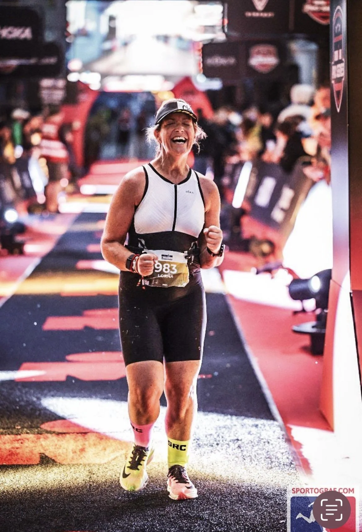 A woman running a race with a victory smile, wearing a white and black athletic outfit, a race bib number 983, and bright yellow and pink running shoes in an indoor or covered running event.