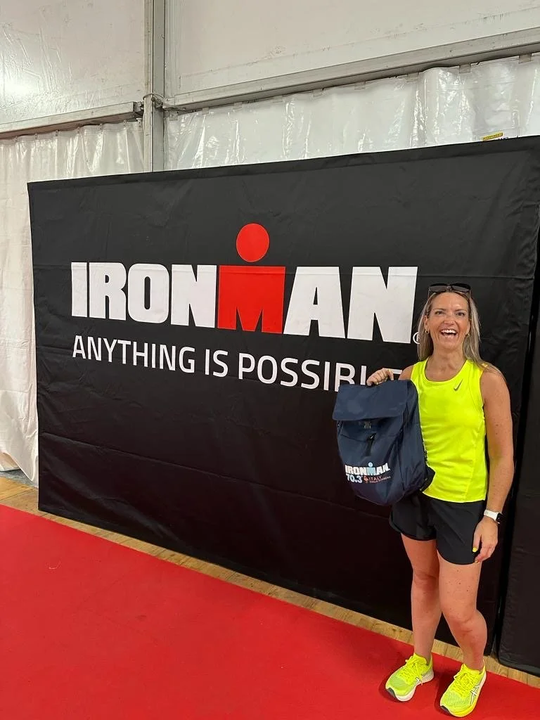 Smiling woman in yellow athletic shirt and black shorts standing next to a black backdrop with 'IRONMAN' logo and tagline, holding a blue gift bag, inside a tent with a red carpet.