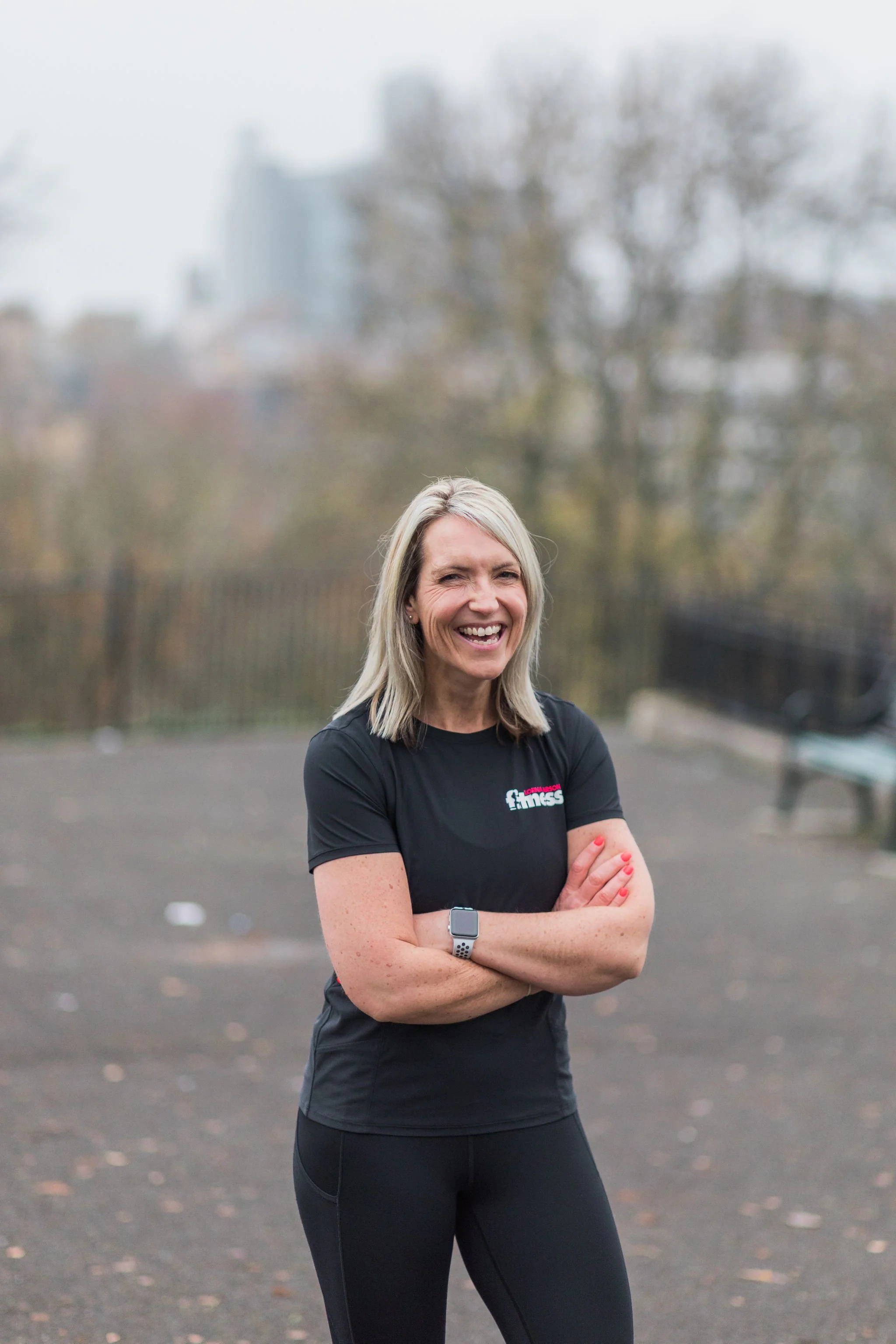 A smiling woman with blonde hair, wearing a black fitness T-shirt and black leggings, standing outdoors with her arms crossed. Background features trees, a bench, and a blurred cityscape.