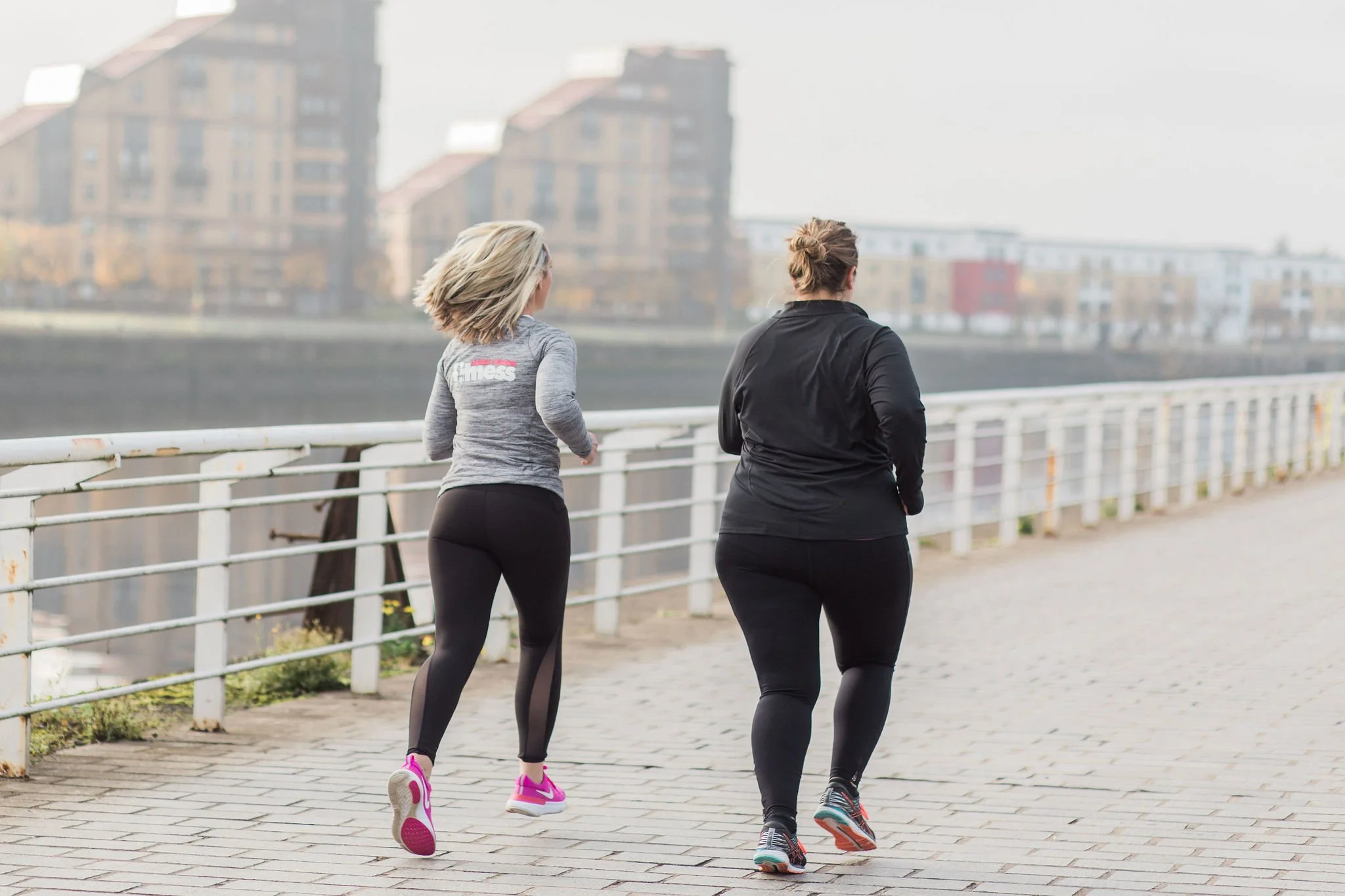 Two women jogging on a paved pathway next to a white railing by a river, with modern buildings in the background.