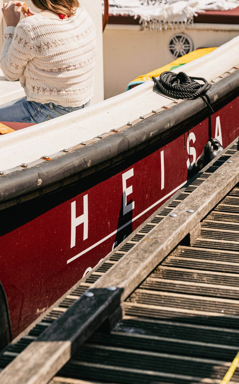 Een close-up van de rode Heisa boot met aan de zijkant het logo, een stuk hout van de kade en links in beeld de rug van iemand die aan boord zit.