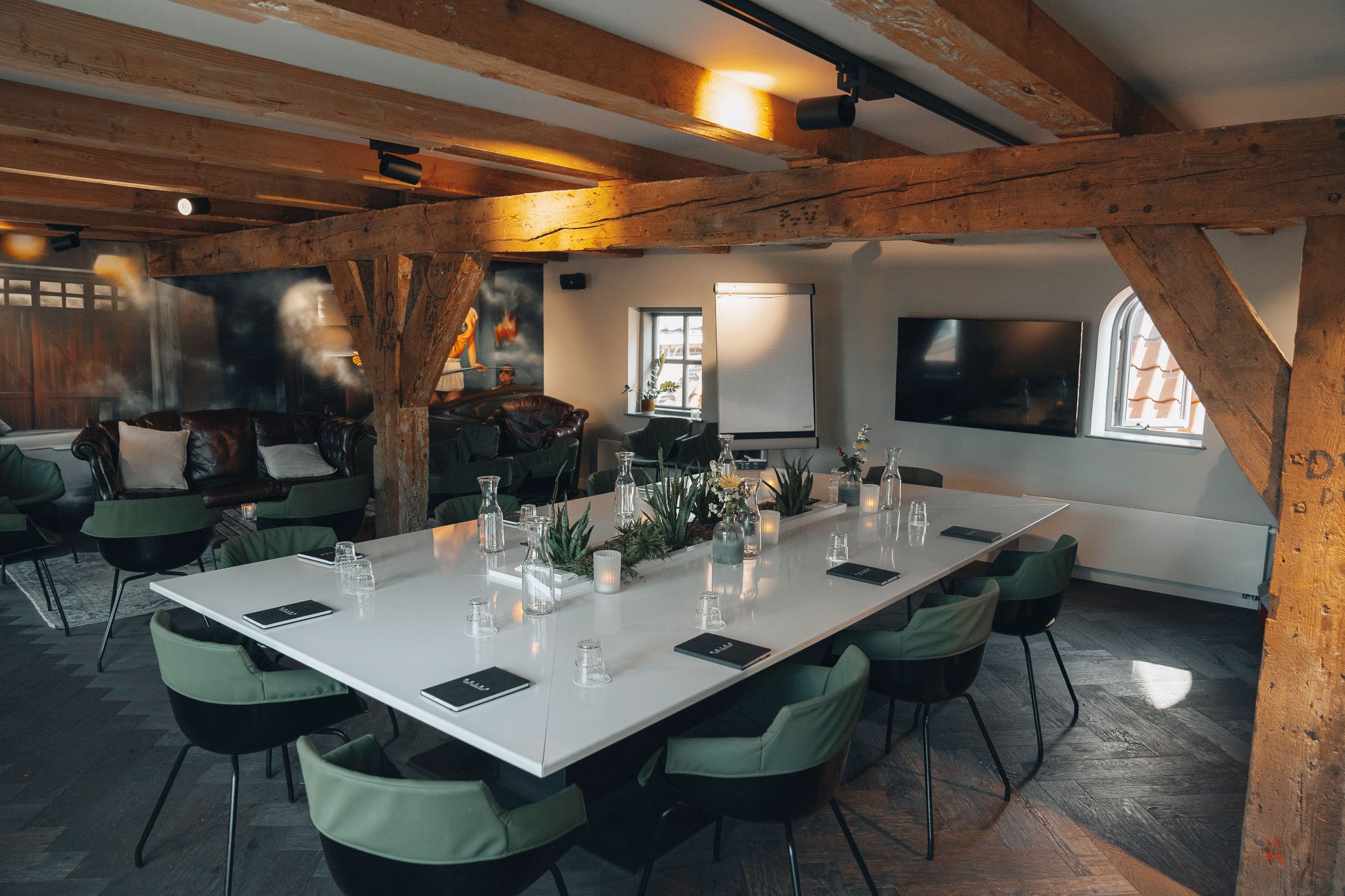 Een gezellige vergaderzaal met een witte tafel, groene stoelen, in een ruimte die gedecoreerd is met kaarsen, planten en muurschilderingen.