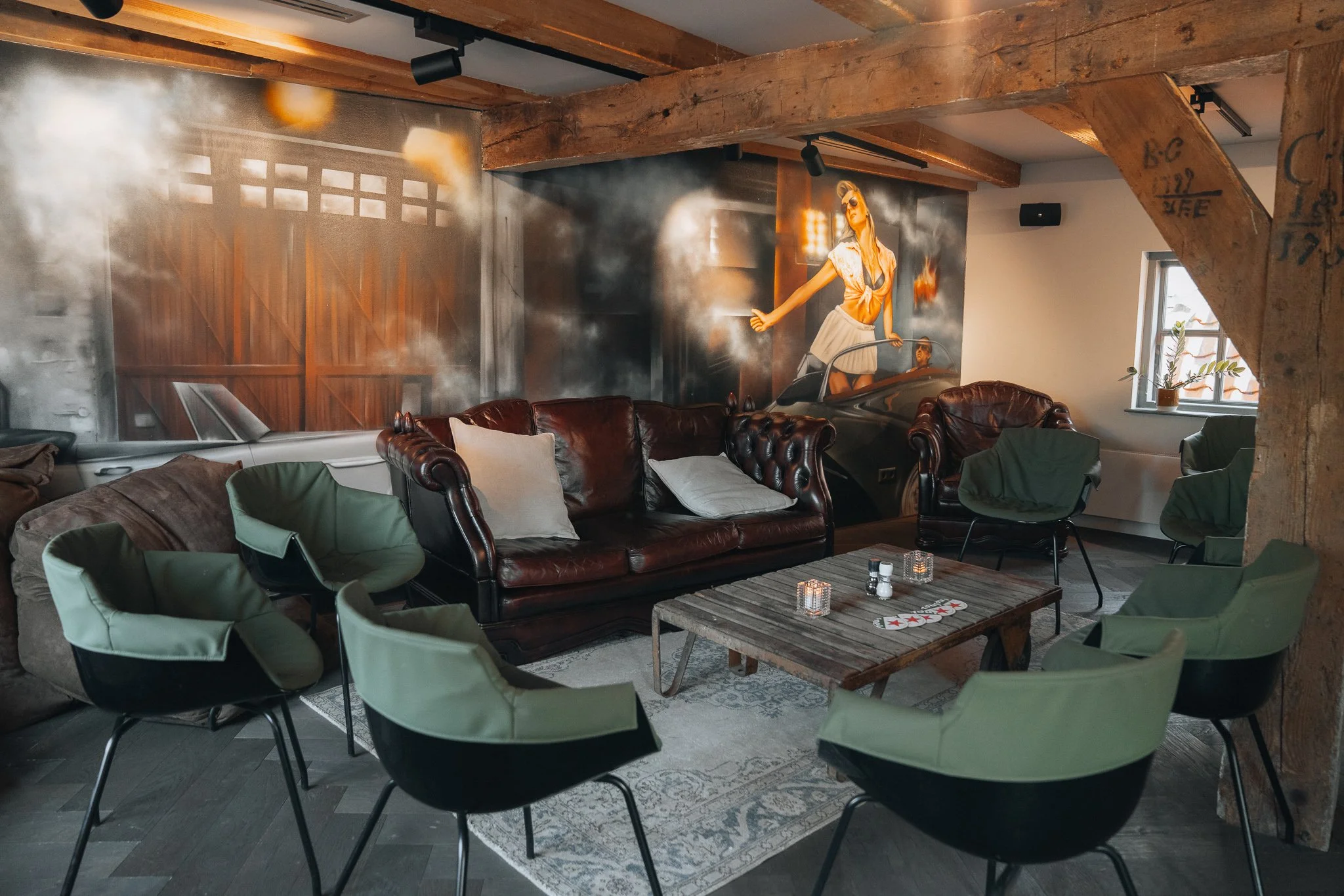 Een ruimte met robuuste houten balken tegen het plafond en groene stoelen die rondom een lage koffietafel staan.