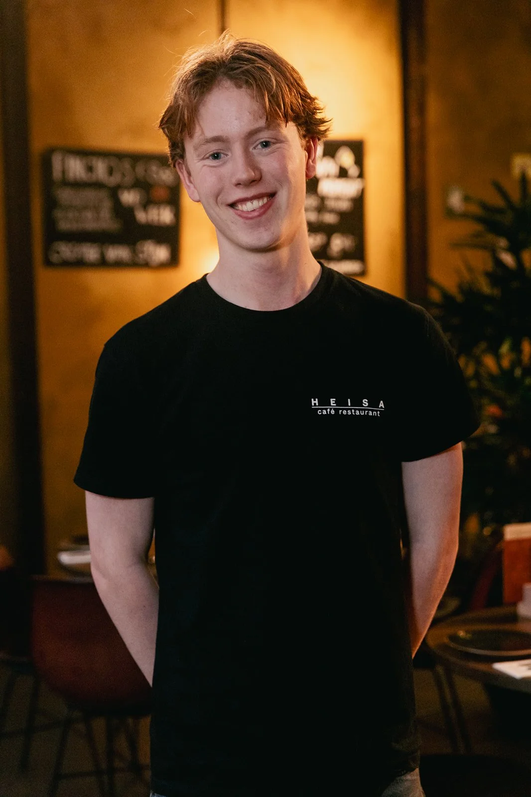Een jongeman staat in een sfeervol restaurant en lacht naar de camera. Hij draagt een zwart T-shirt met daarop het logo van Heisa.