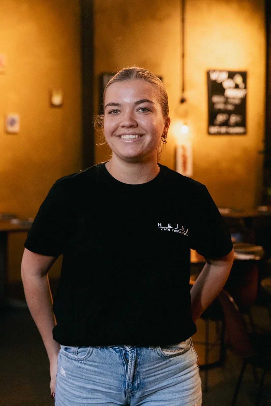 Een personeelslid van café restaurant Heisa lacht naar de camera in het sfeervol verlichte café.
