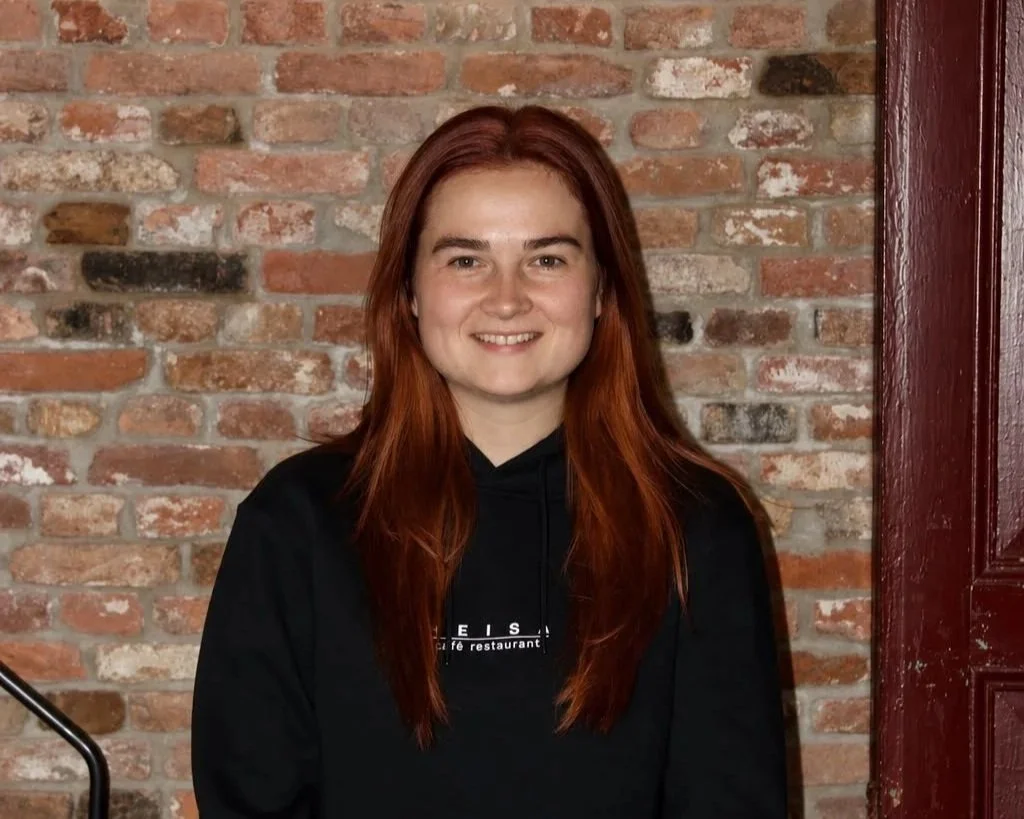Een vrouw met paardenstaart heeft een hand in de zij en lacht naar de camera in een zwart T-shirt met daarop het logo van café restaurant Heisa.