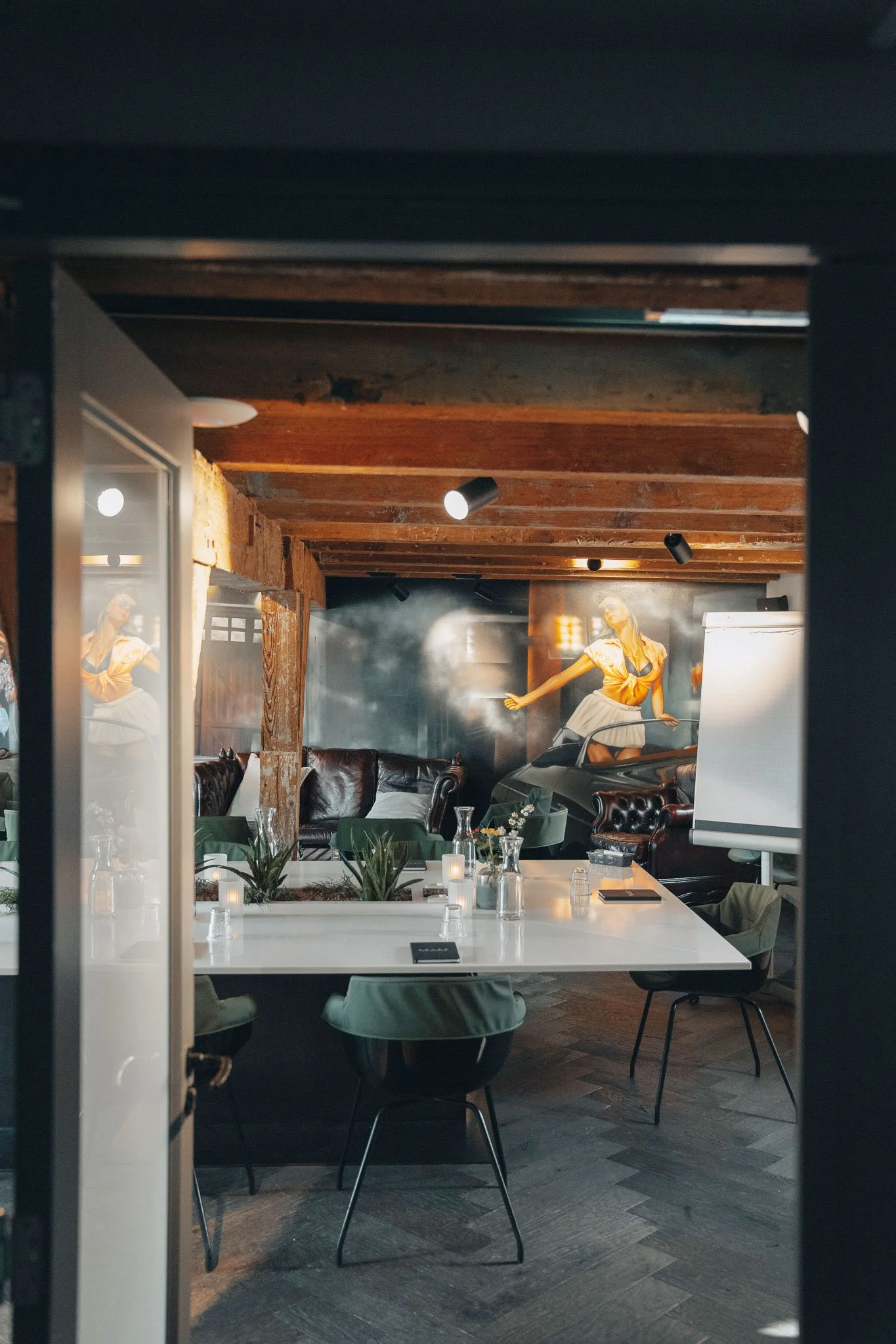 Een gezellige vergaderzaal met een witte tafel, groene stoelen, in een ruimte die gedecoreerd is met kaarsen, planten en muurschilderingen.