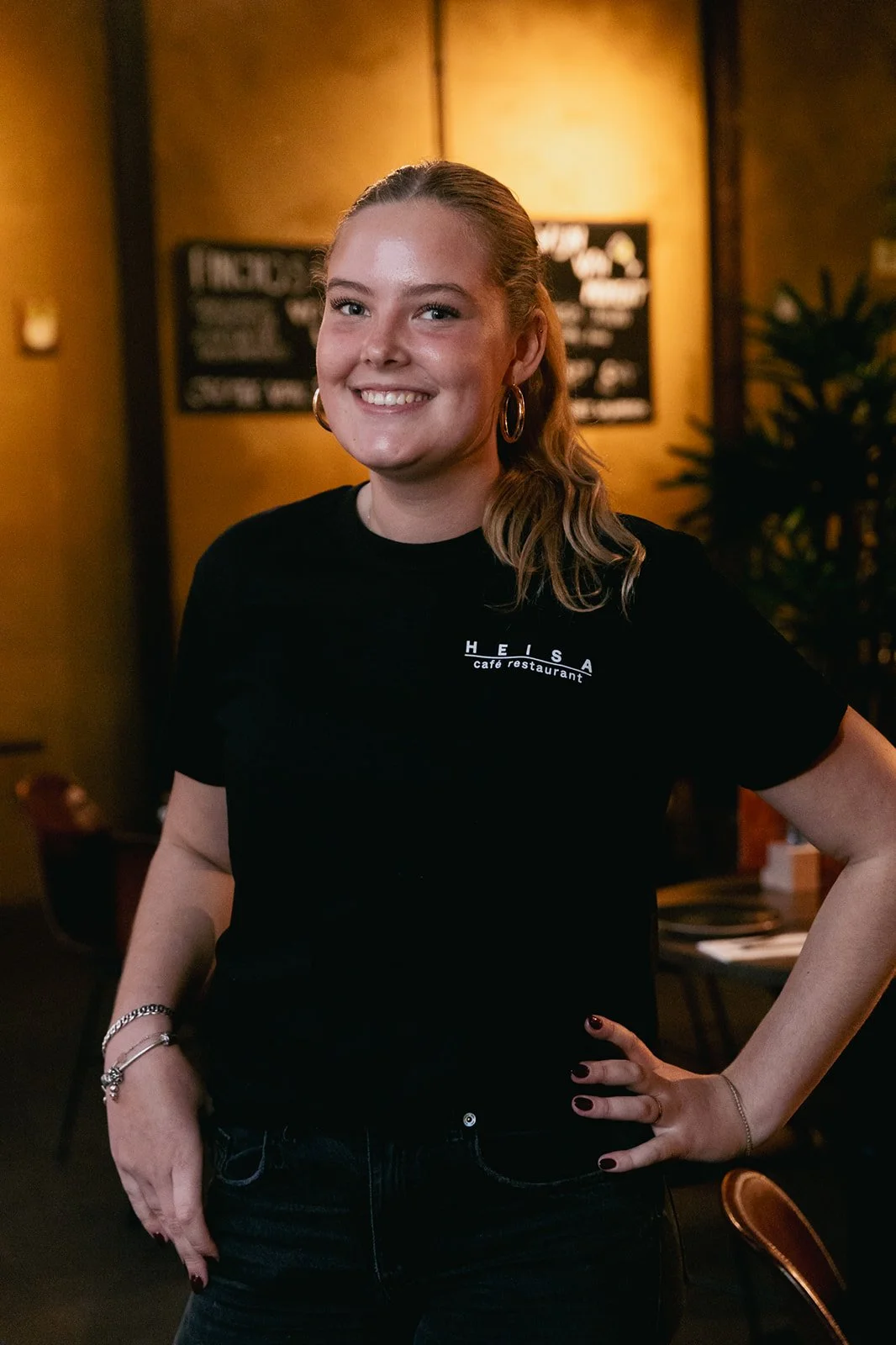 Een personeelslid met blond haar lacht naar de camera en draagt een zwart T-shirt met daarop het logo van Heisa.