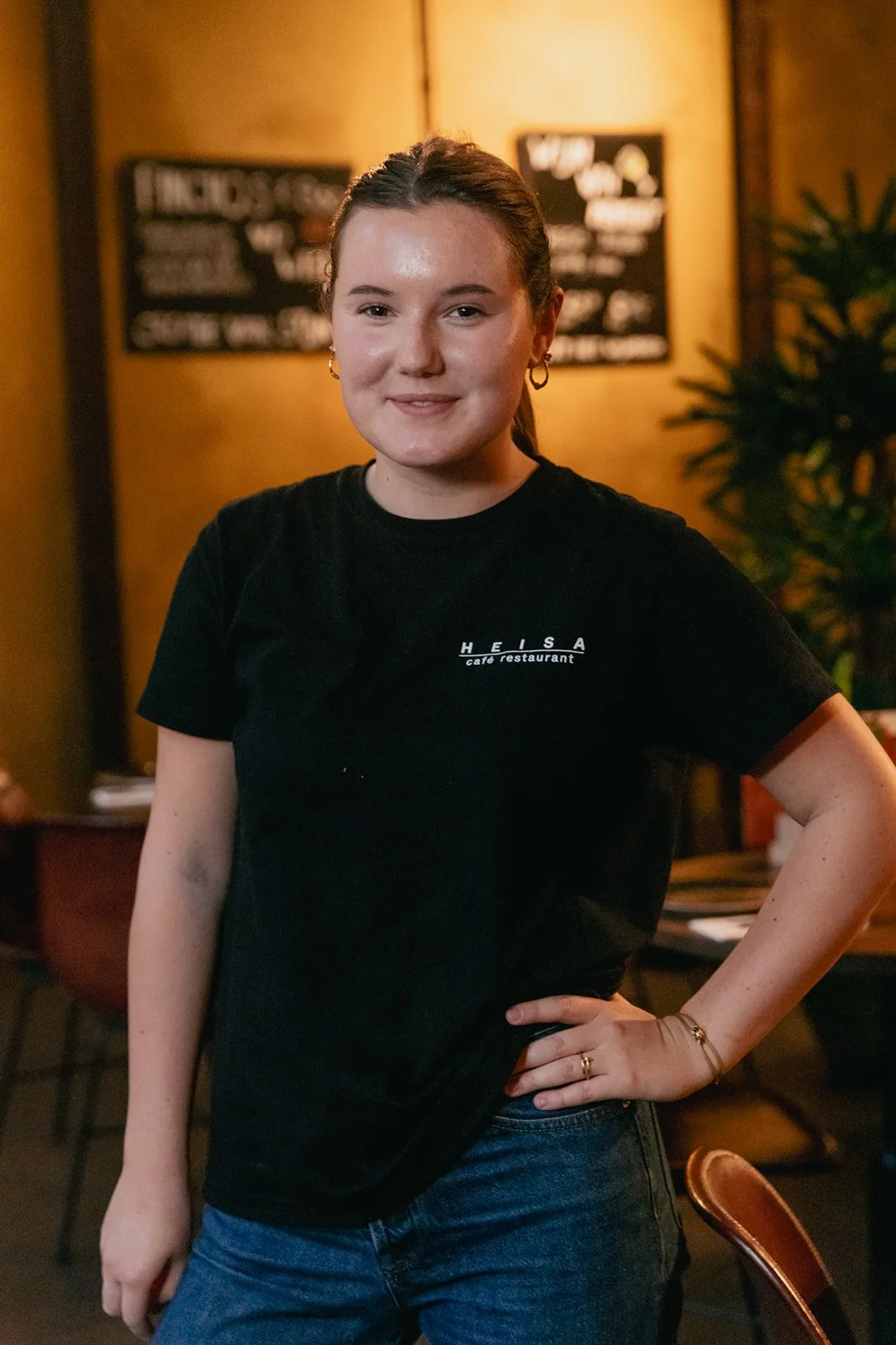 Een vrouw staat in een sfeervol restaurant met een hand in haar zij en lacht naar de camera. Ze draagt een zwart T-shirt met daarop het logo van Heisa.