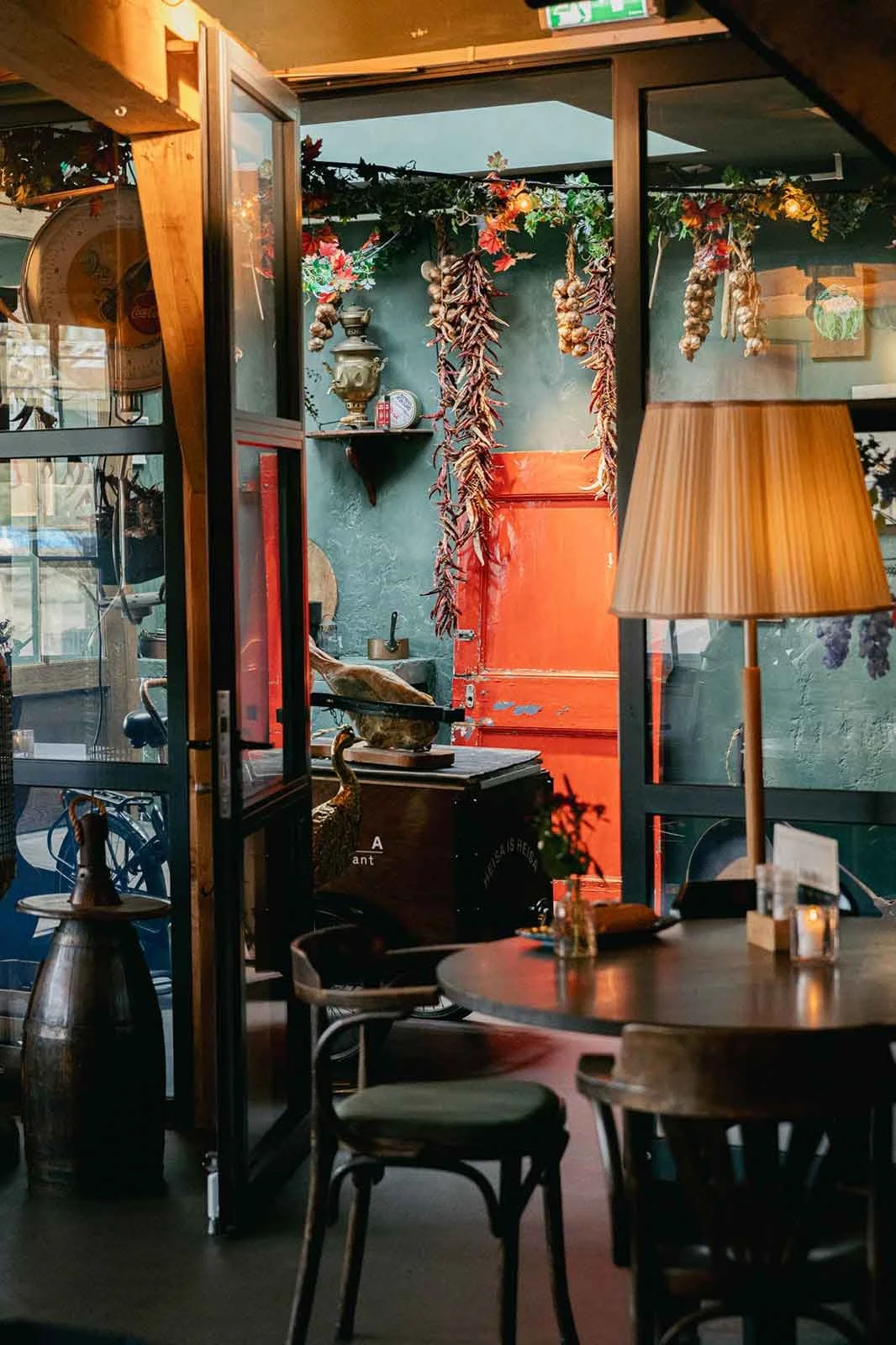 Interieur van een gezellig restaurant of café met een ronde tafel, houten stoelen en een staande lamp. Op de achtergrond is een deur te zien met hangende gedroogde knoflook en kruiden, decoratieve voorwerpen op een plank en warme verlichting.