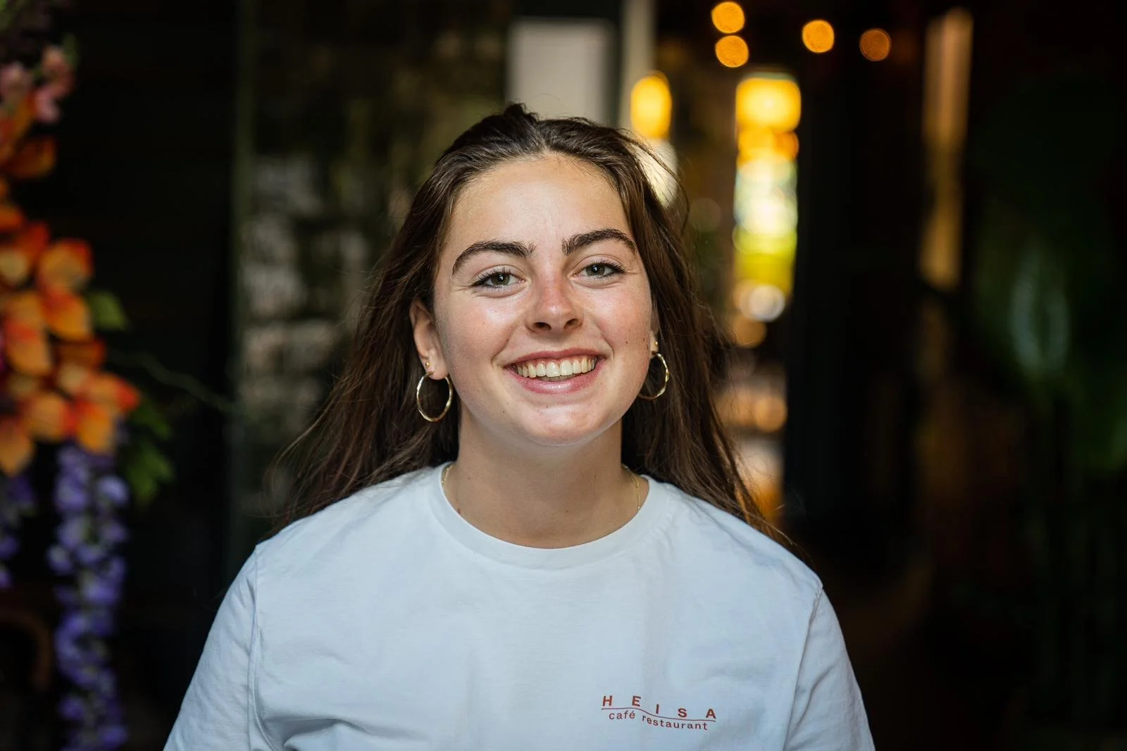 Een personeelslid van Heisa lacht naar de camera in het sfeervol verlichte restaurant. Ze draagt een wit T-shirt waar Heisa op staat.