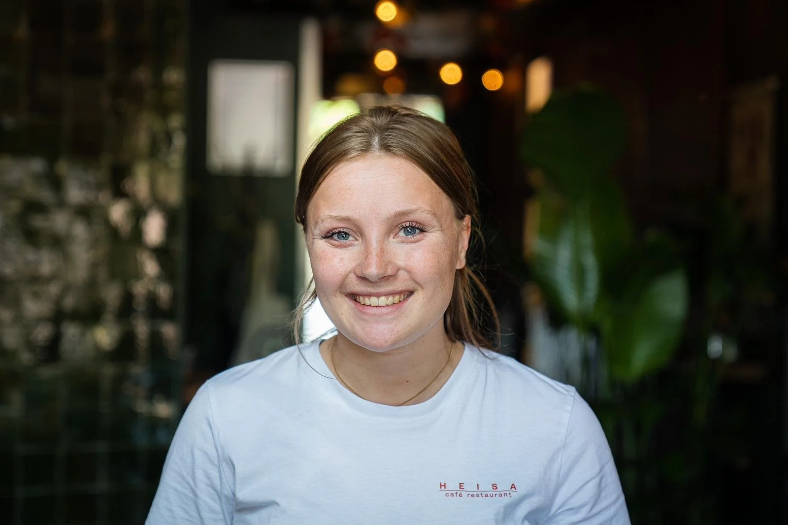 Een vrouw uit de bediening van Heisa lacht naar de camera in het sfeervol verlichte restaurant. Ze draagt een wit T-shirt waar Heisa op staat