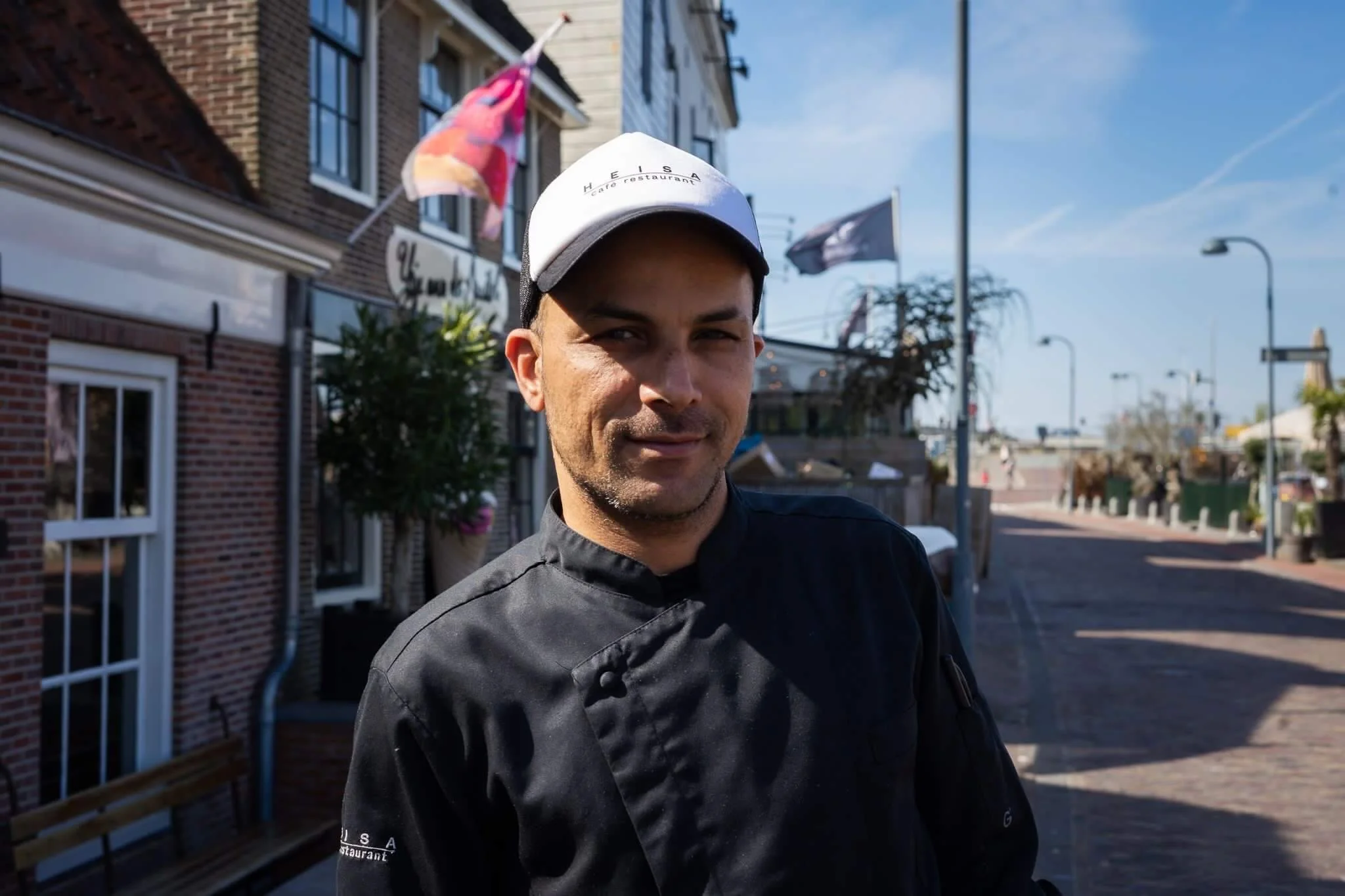 Een man uit het keukenteam van Heisa staat op de stoep van het restaurant en kijkt naar de camera. Hij draagt een witte pet en een zwart kokspak.