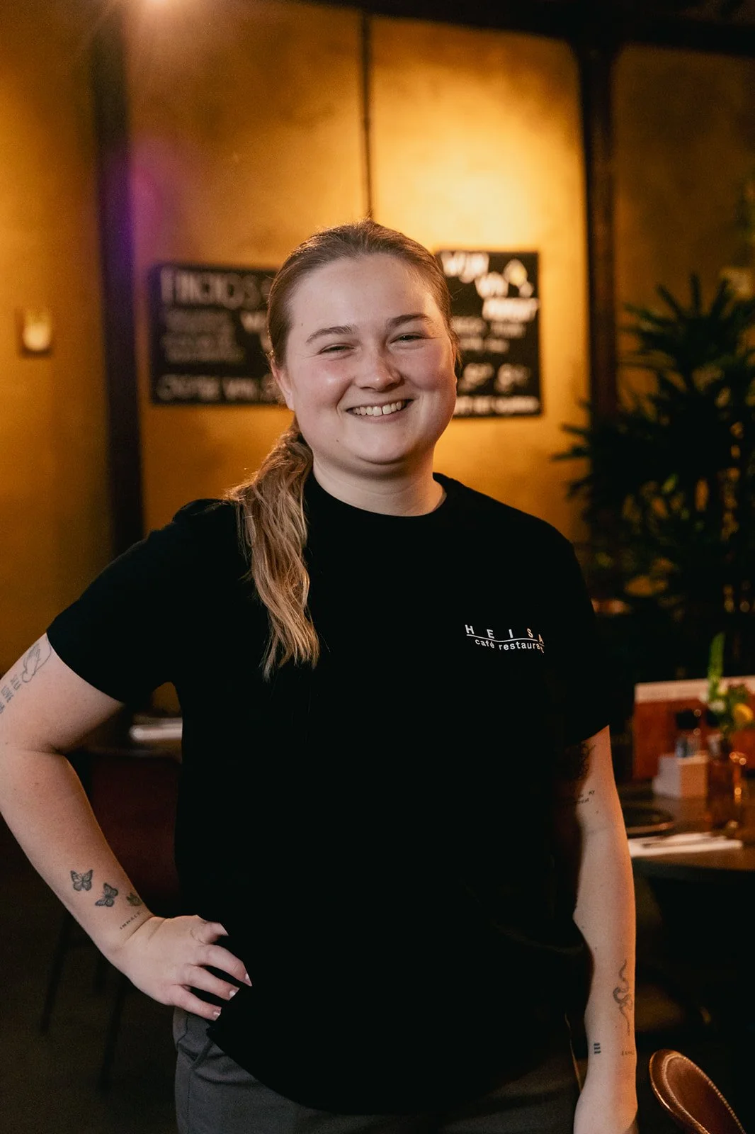 Een vrouw met paardenstaart heeft een hand in de zij en lacht naar de camera in een zwart T-shirt met daarop het logo van café restaurant Heisa.