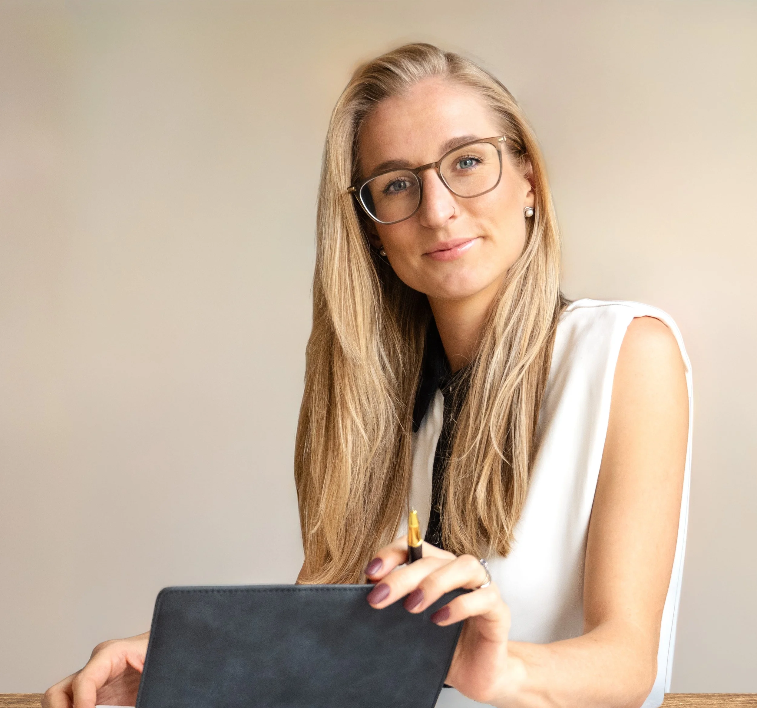 A woman with long blonde hair, glasses, and earrings sitting at a desk, holding a pen and a closed notebook, looking at the camera with a slight smile.