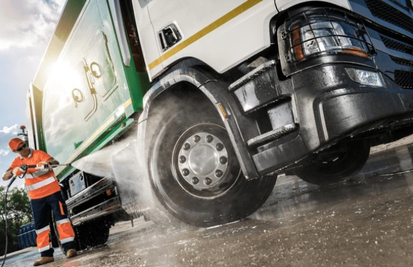 Worker in orange safety gear pressure washing the front wheel of a large truck with a green and white trailer on a wet concrete surface.