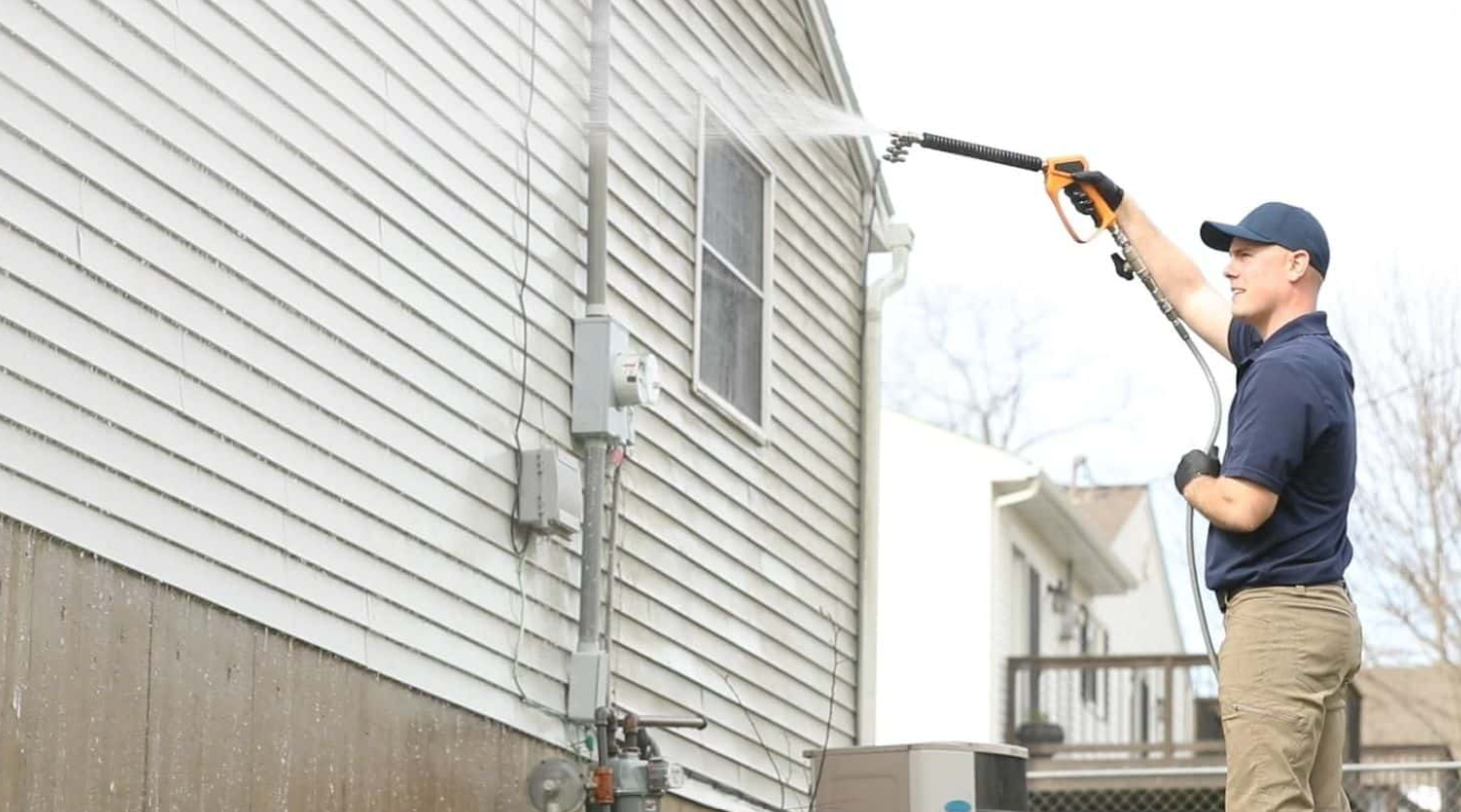 Man in navy shirt and khaki pants pressure washing the exterior siding of a house.