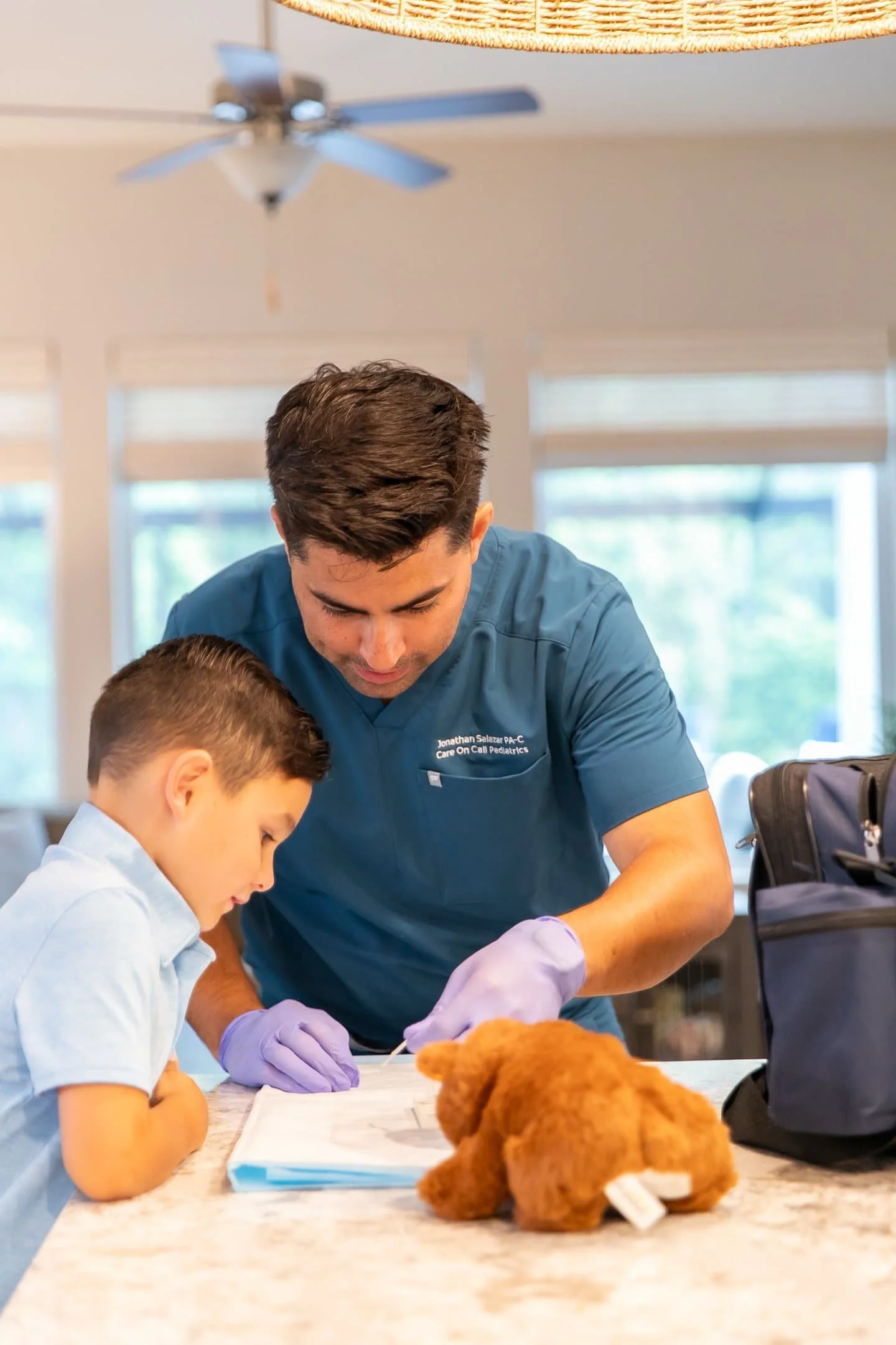 A healthcare professional wearing purple gloves and a blue uniform assisting a young boy with a medical task at a table, with a teddy bear and a black bag nearby in a bright room.