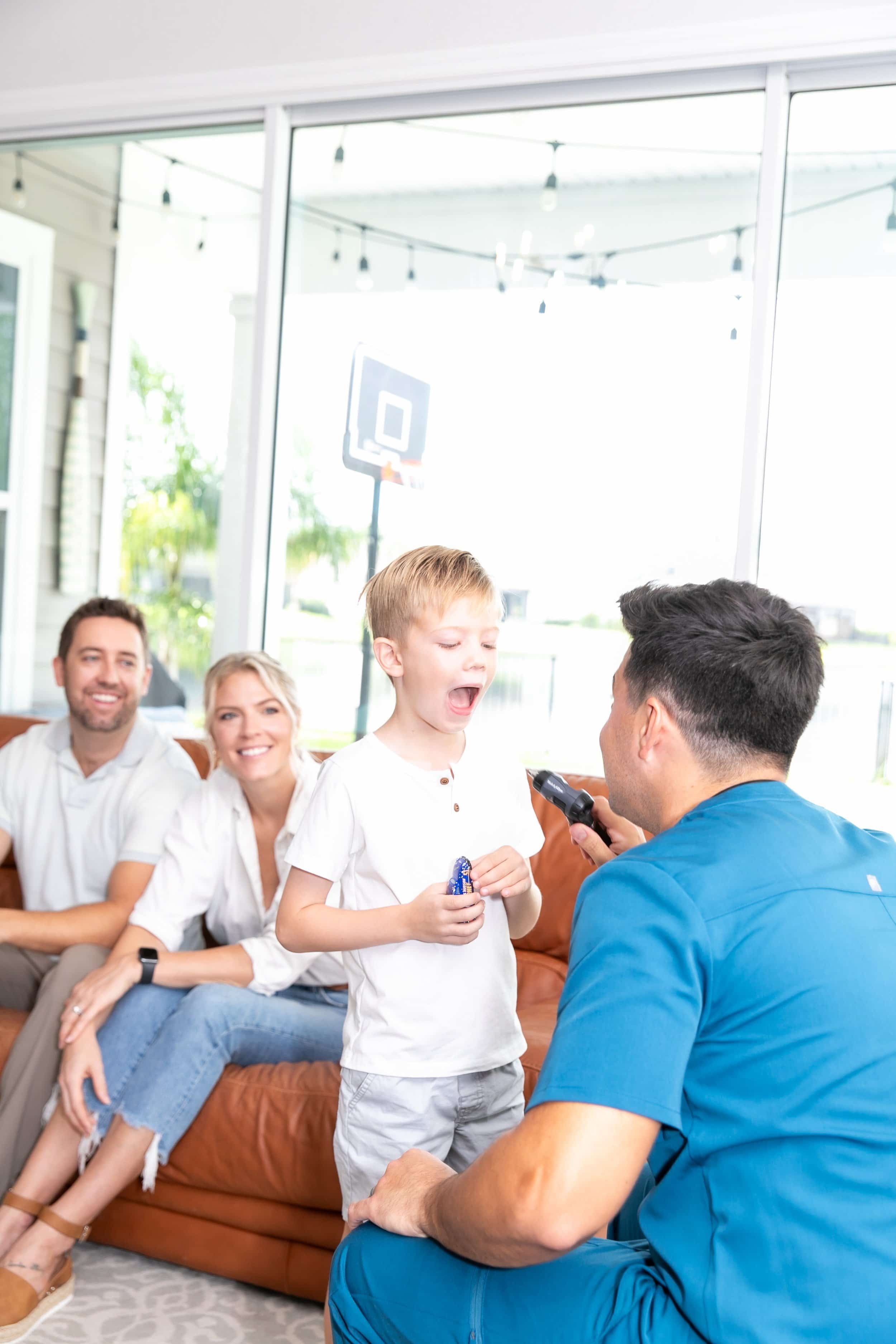 A young boy with a white shirt is crying while holding a toy, as a man in blue scrubs appears to be using a medical device on him. Family members sit on a brown sofa behind them, smiling and watching the scene, in a brightly lit room with large windows.