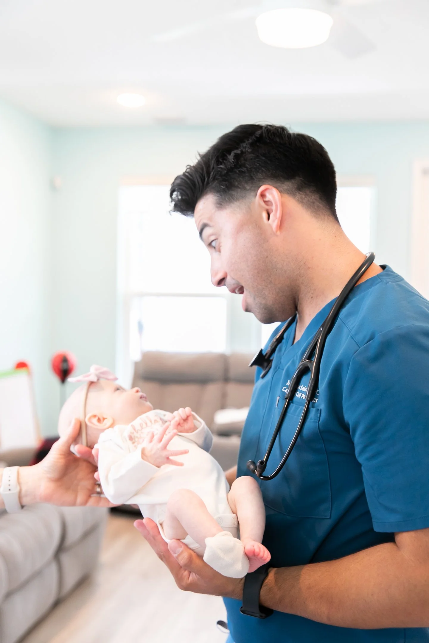 A smiling doctor in blue scrubs holds a baby, gently talking to her in a bright, cozy room, showcasing caring pediatric care.