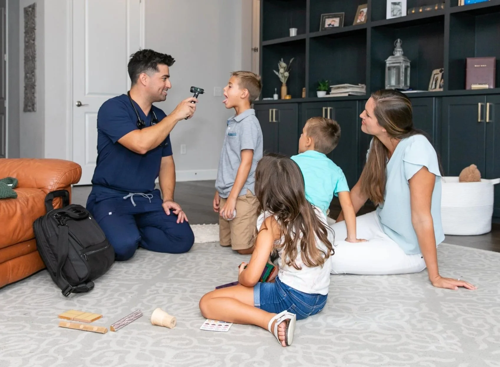 A House Call Pediatrics doctor examines a young boy’s throat with an otoscope while his family watches in the living room.