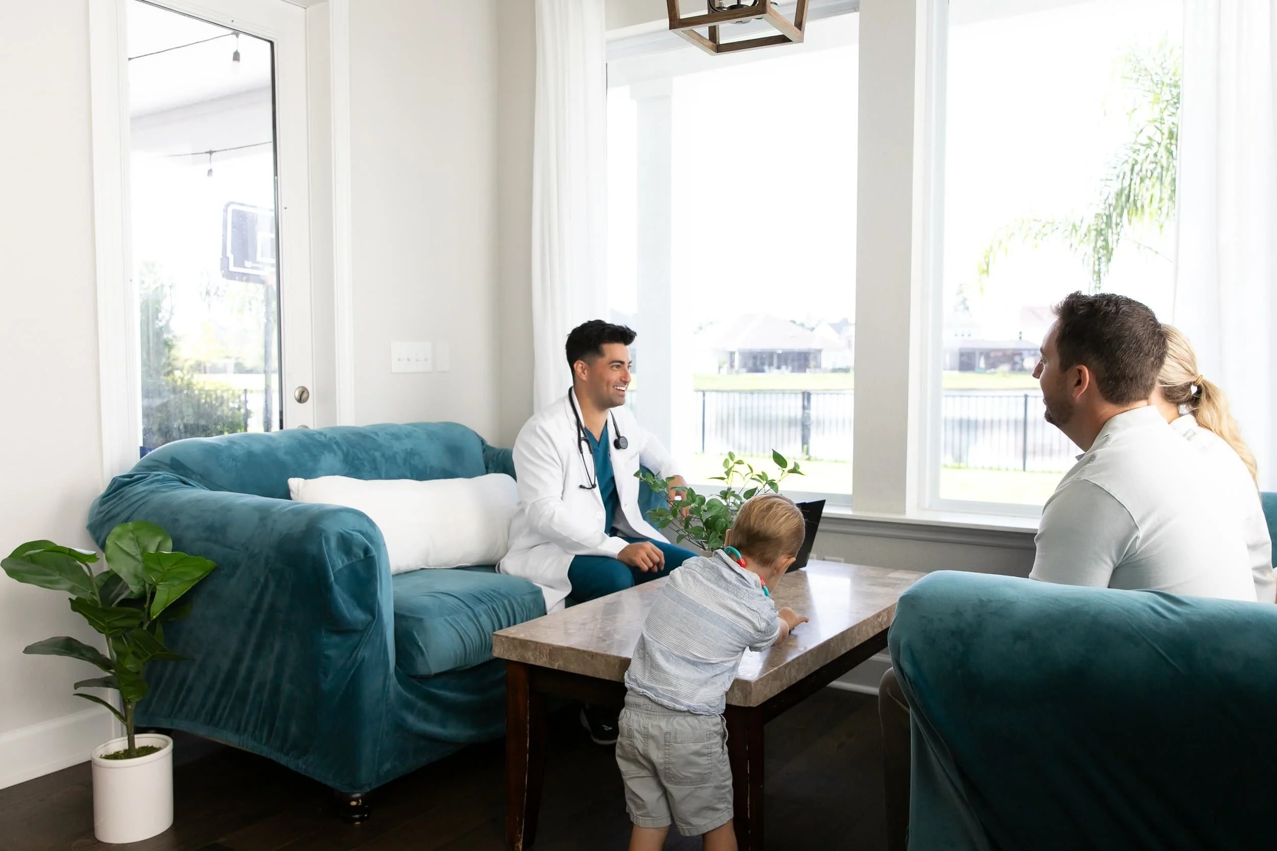 A young male doctor with a stethoscope around his neck sitting on a teal sofa, smiling, during a conversation with a man and a young boy in a bright living room with large windows and green plants.