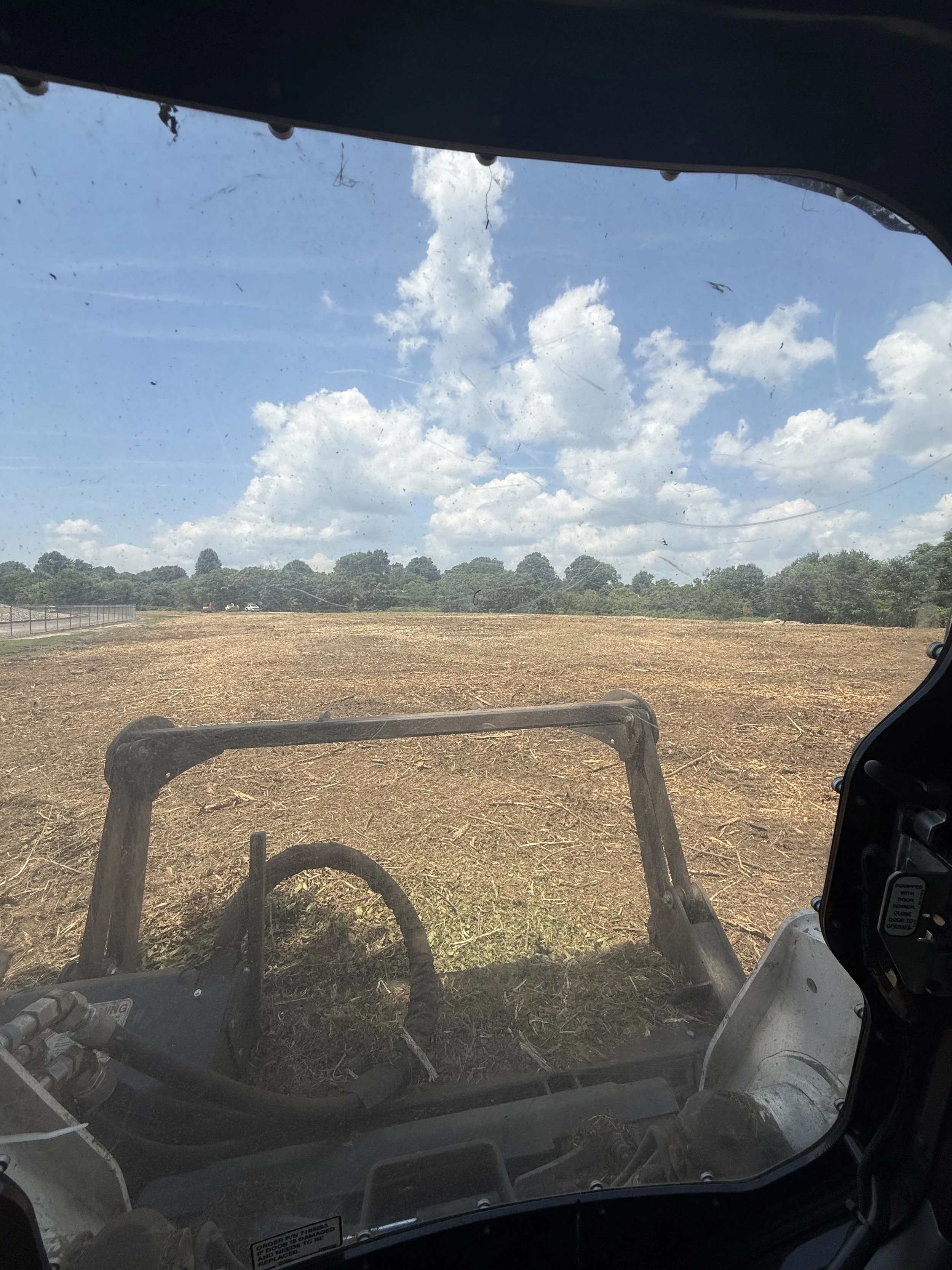 View from inside a construction or farming vehicle looking out over a dirt field with a line of trees in the distance and a partly cloudy sky.