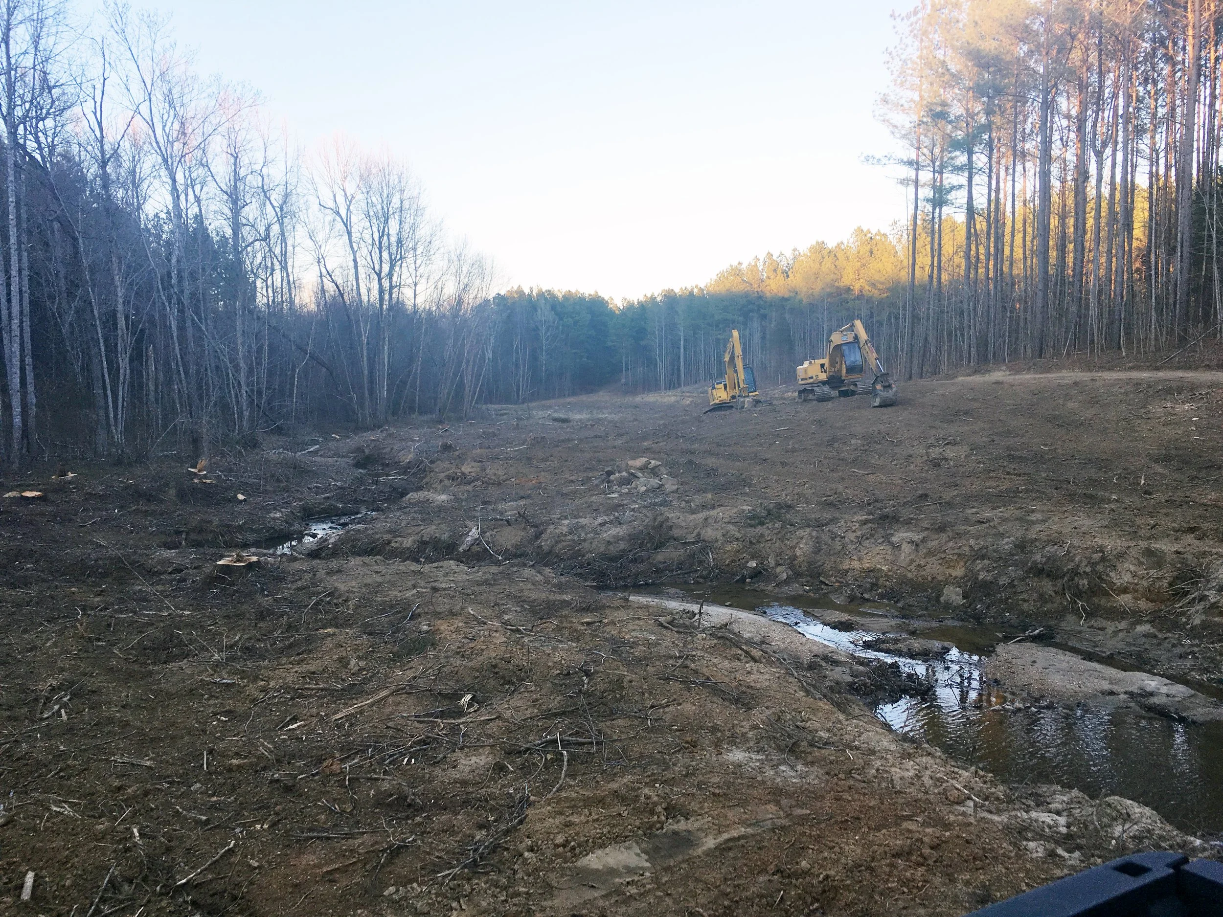 Construction equipment on a cleared area of land next to a forest, with two excavators working and a small stream running through the dirt.