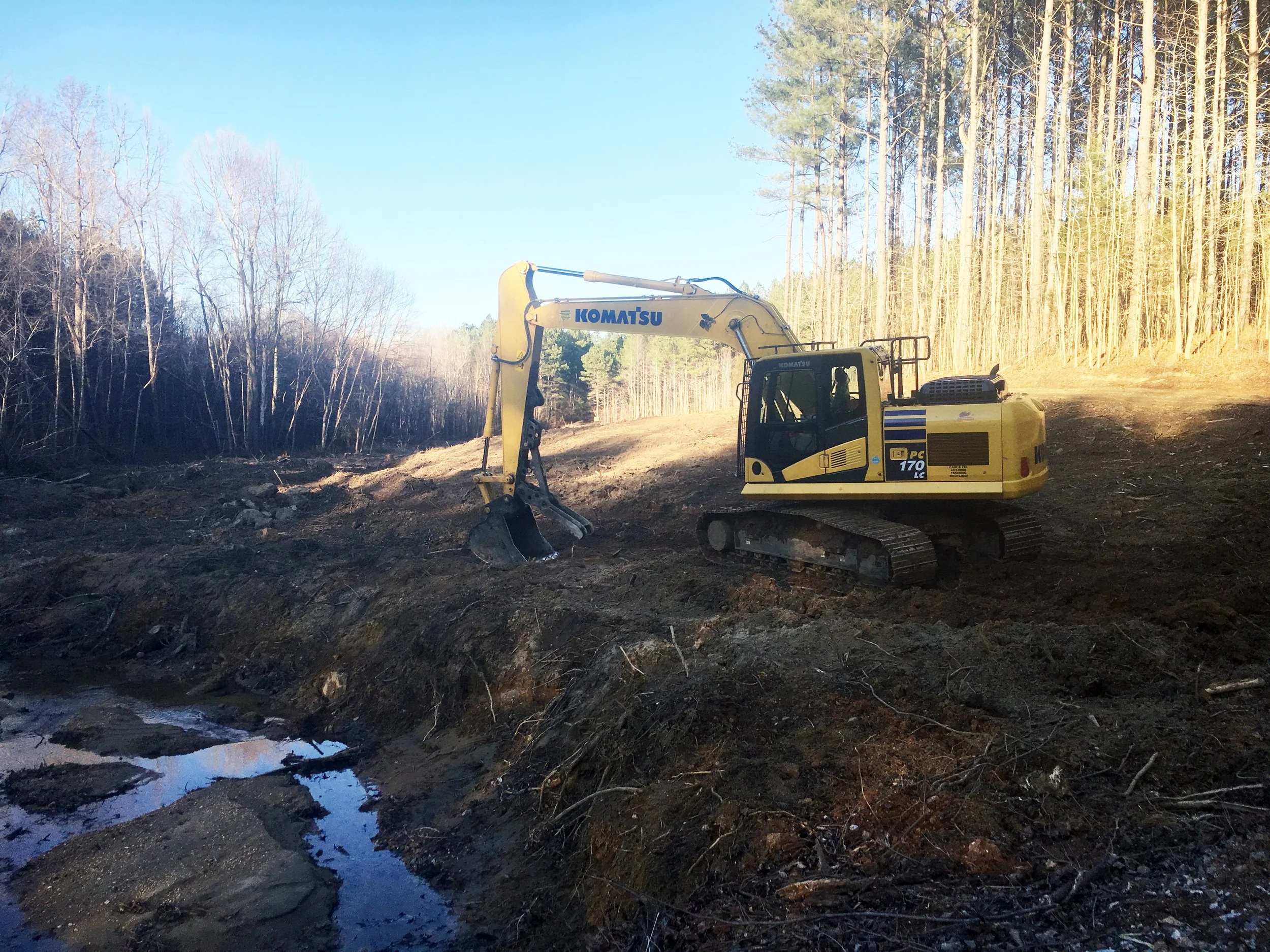 A yellow Komatsu excavator working on a dirt area near a small stream, with a forest of leafless and evergreen trees in the background during daylight.