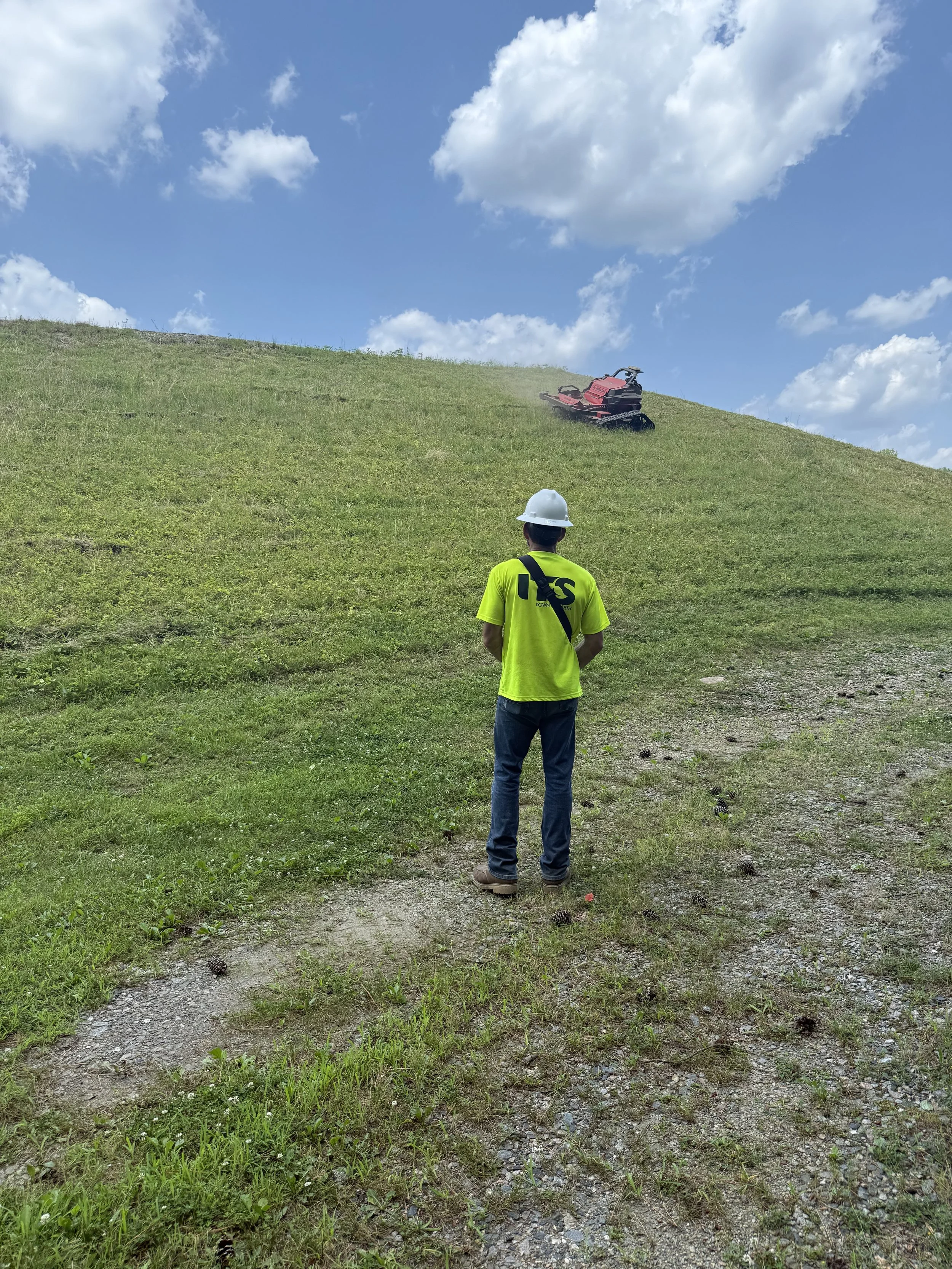 A man wearing a white safety helmet and a bright yellow shirt stands on a grassy hill, looking at a red tracked vehicle driven by a dust cloud at the top of the hill under a partly cloudy sky.