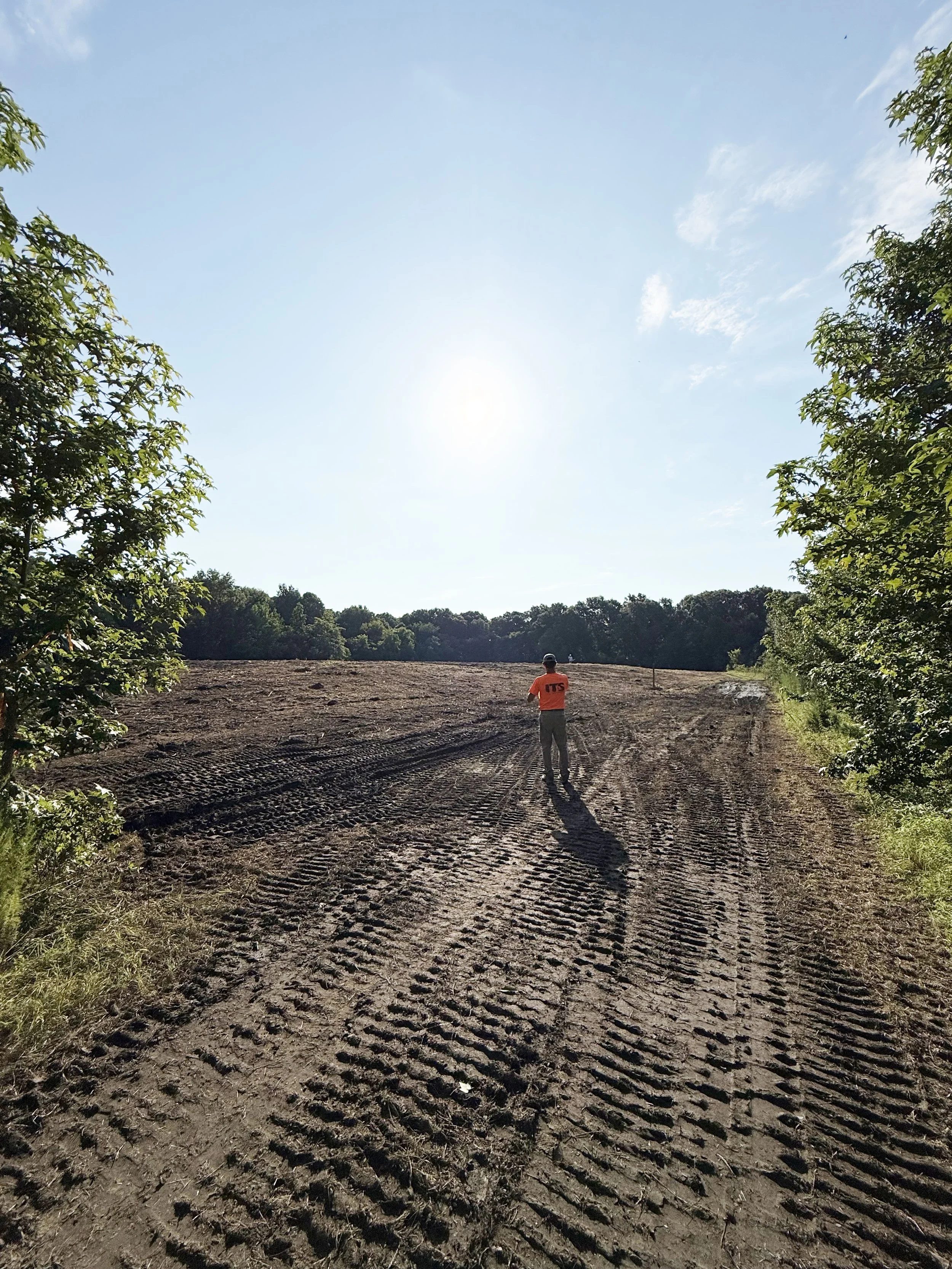 A person standing on a dirt path or construction site, wearing an orange shirt with 'TS' on the back, surrounded by trees and under a sunny blue sky.