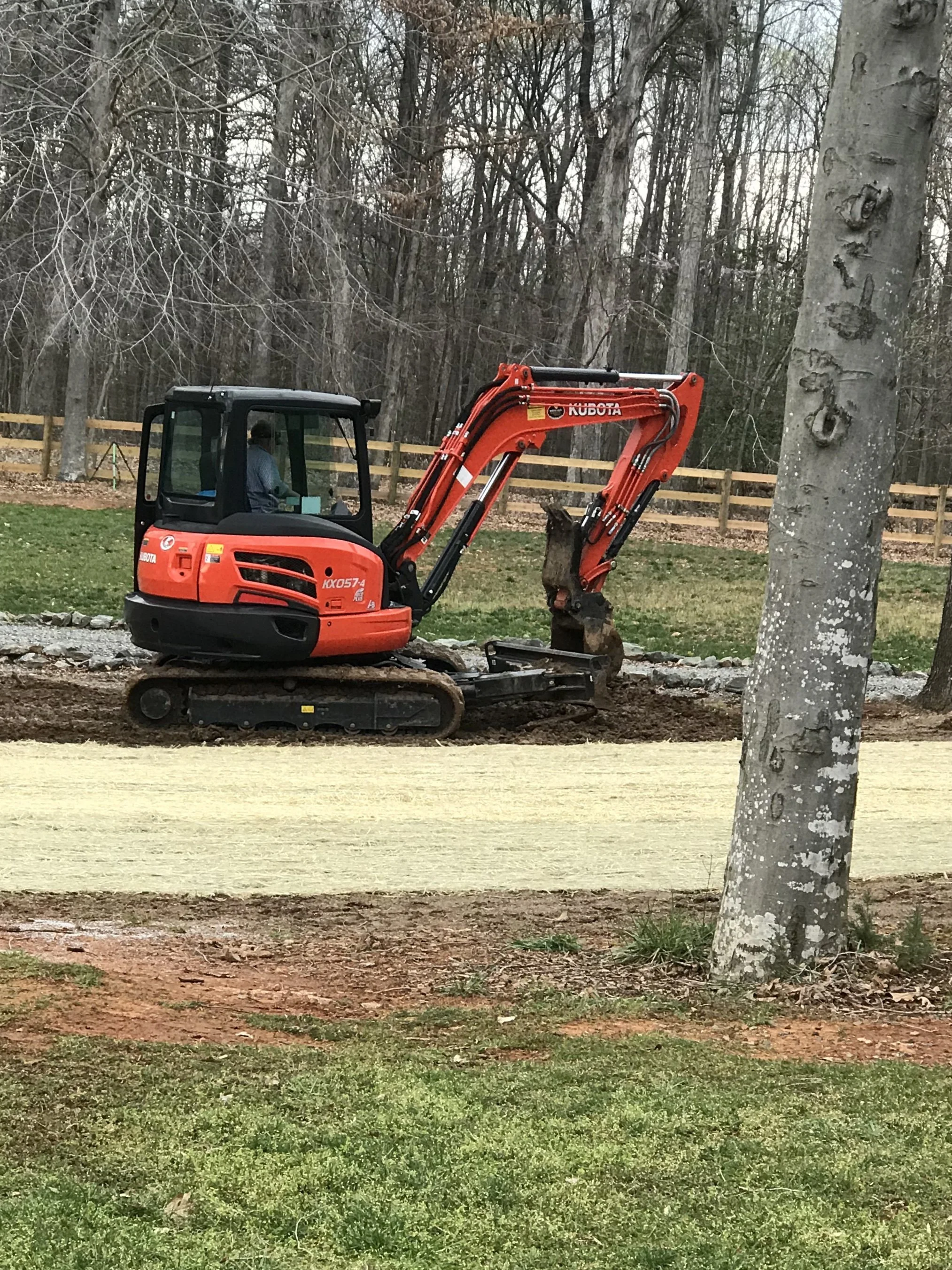 A small orange excavator working on a landscape or garden project with a dirt path and trees in the background.