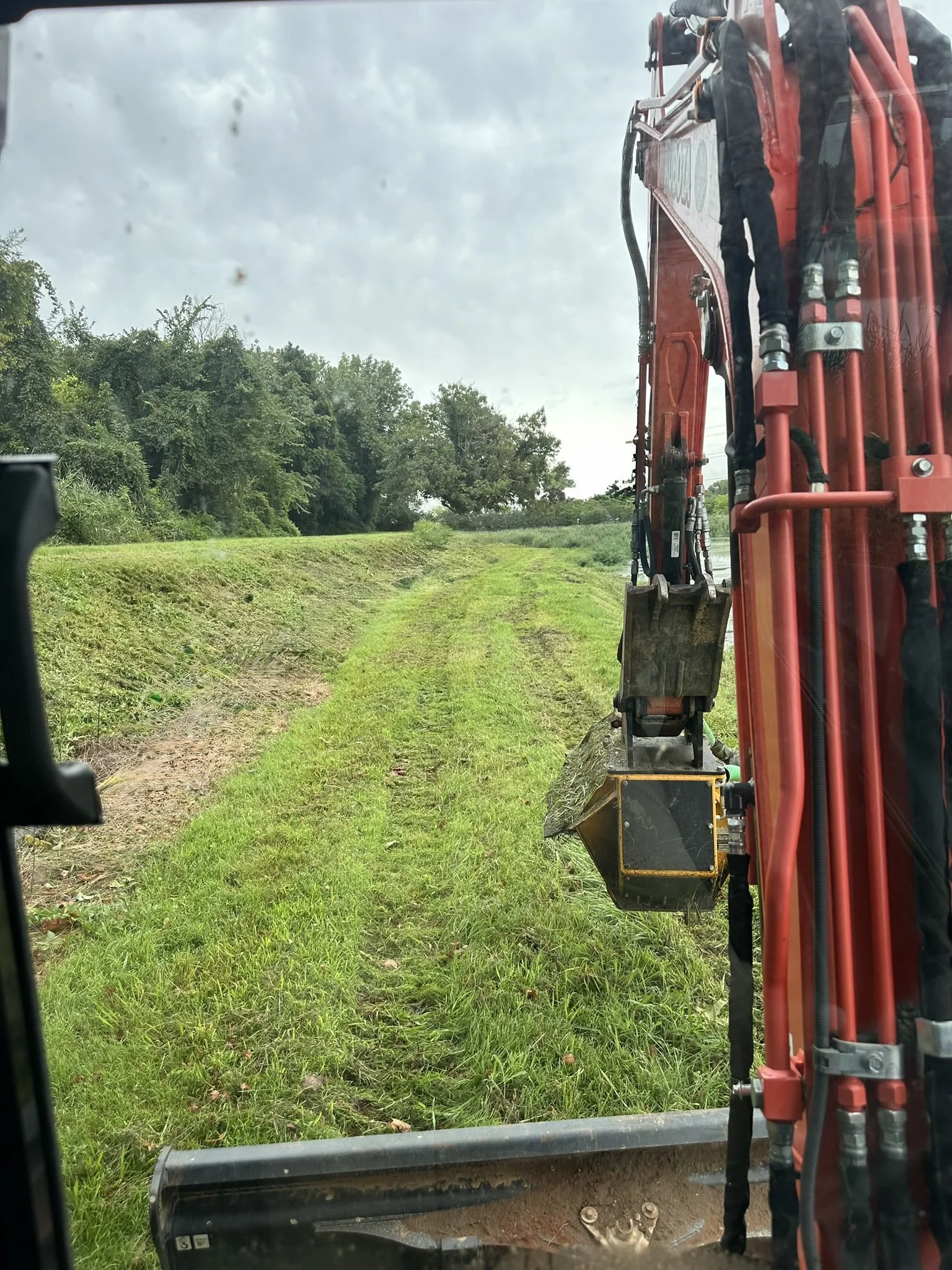 View from inside construction vehicle looking at a grassy, partially mowed path with green trees on left and cloudy sky overhead.